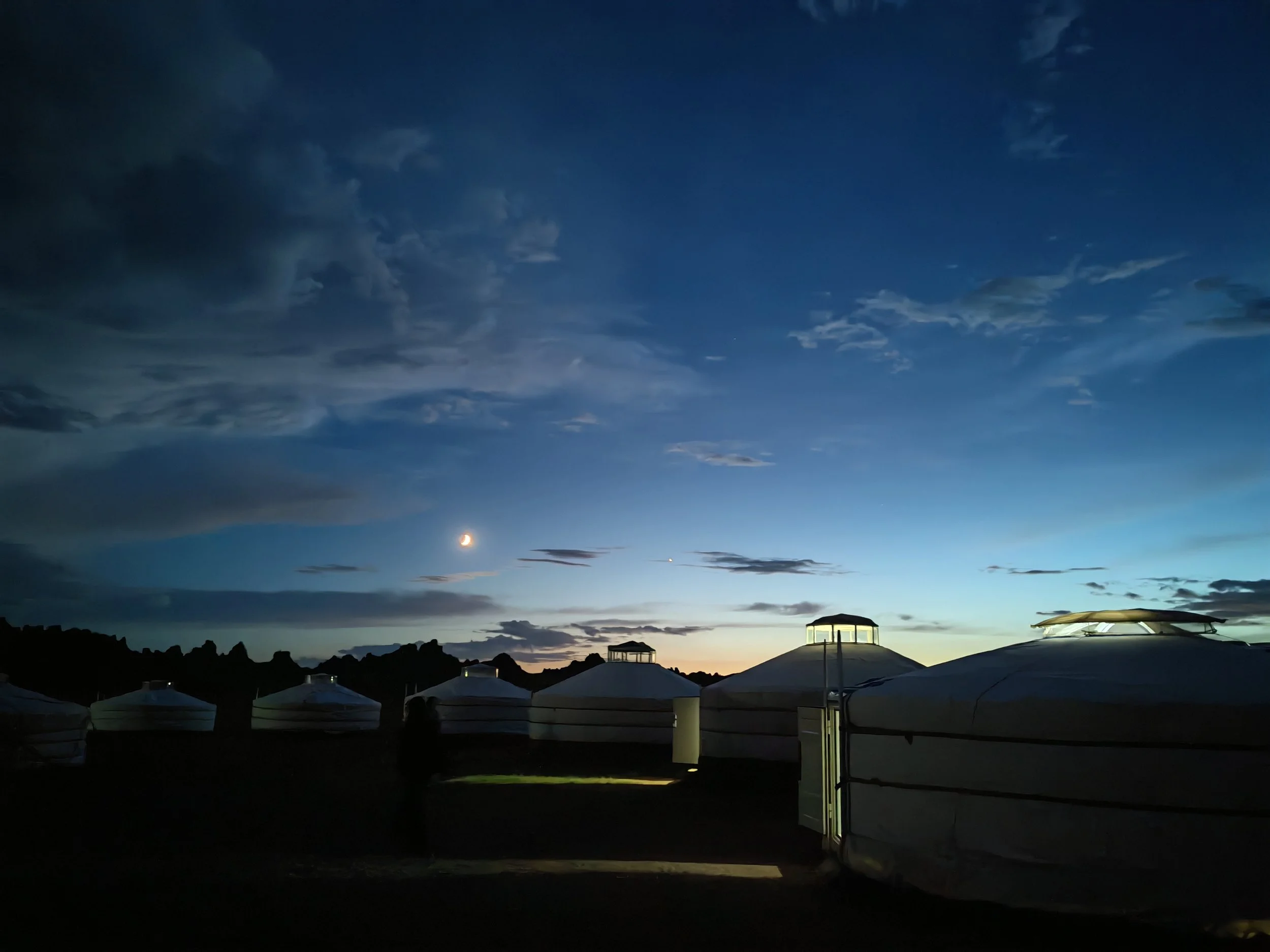 A nighttime scene with a crescent moon in the darkening sky and some clouds. The horizon shows silhouettes of mountains and several large tents or structures with rounded tops, illuminated from below.