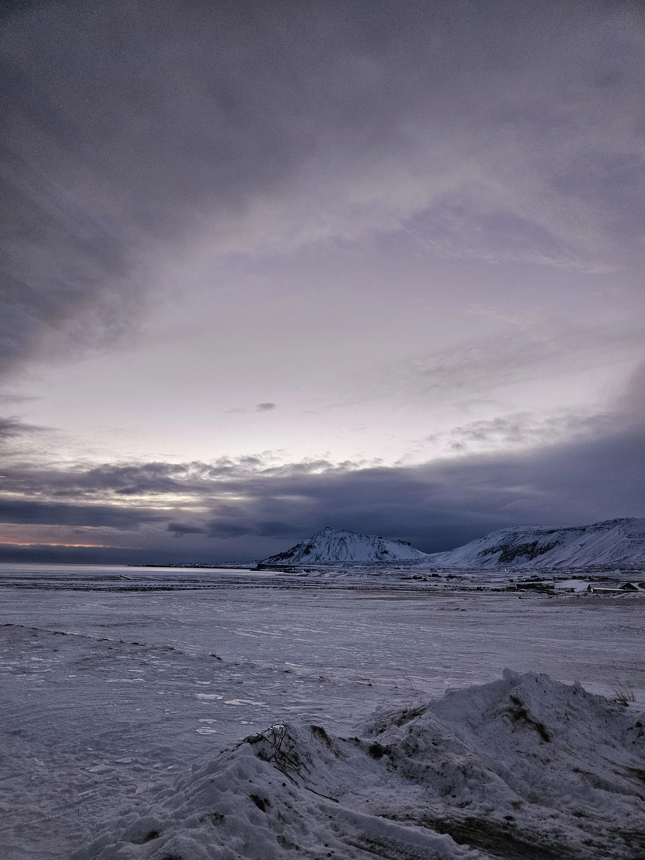 A snowy landscape with mountains in the distance under a cloudy sky at dusk or dawn.