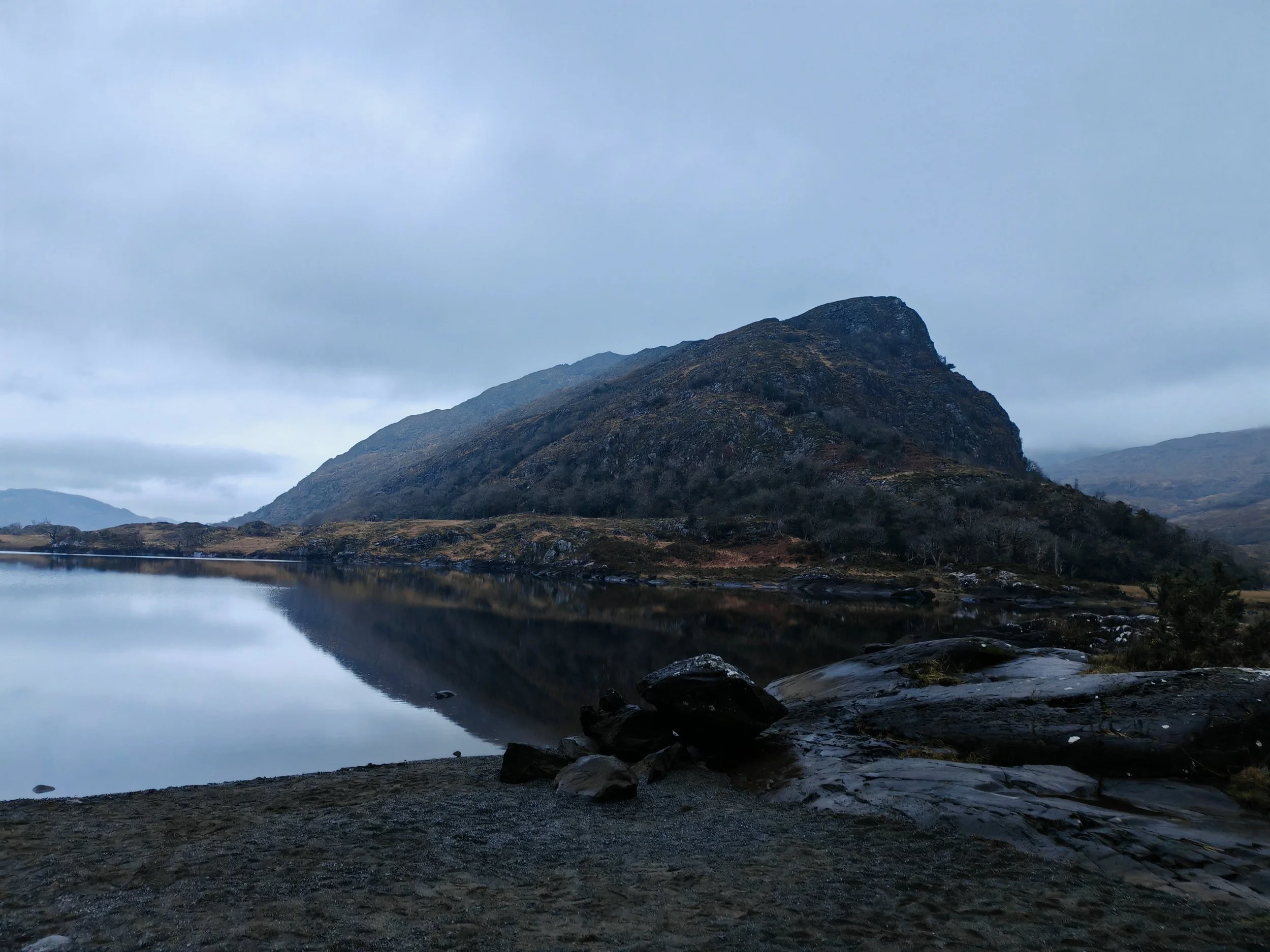 A mountain landscape with a lake in the foreground, reflecting the mountain and sky, under a cloudy sky.