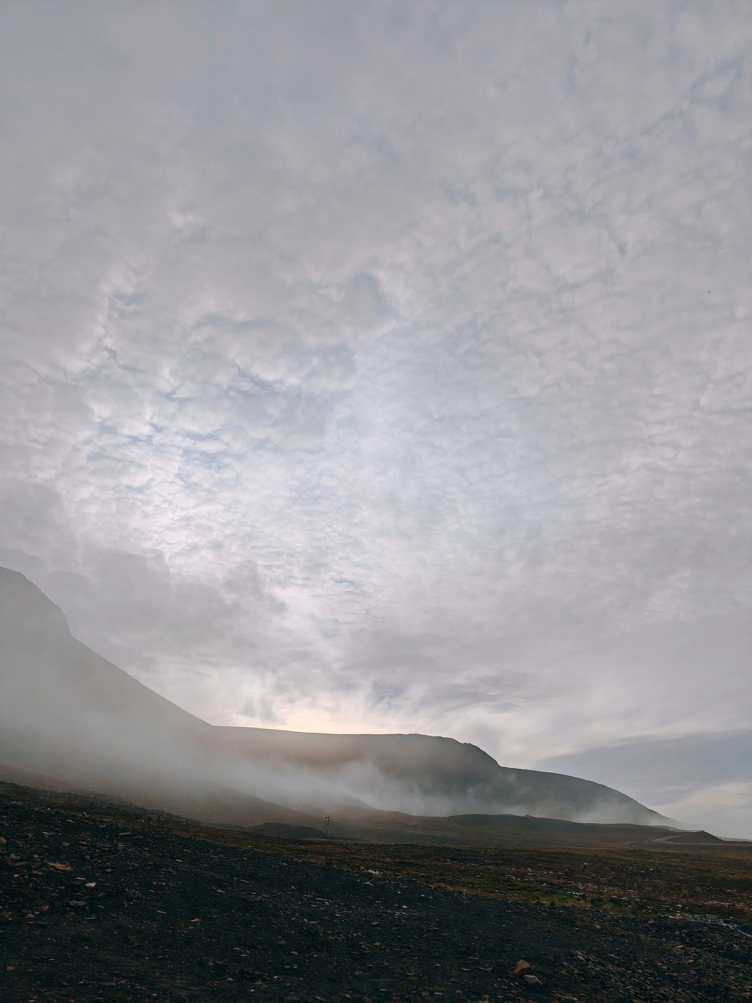 A mountainous landscape under a cloudy sky with dark soil and sparse vegetation in the foreground.