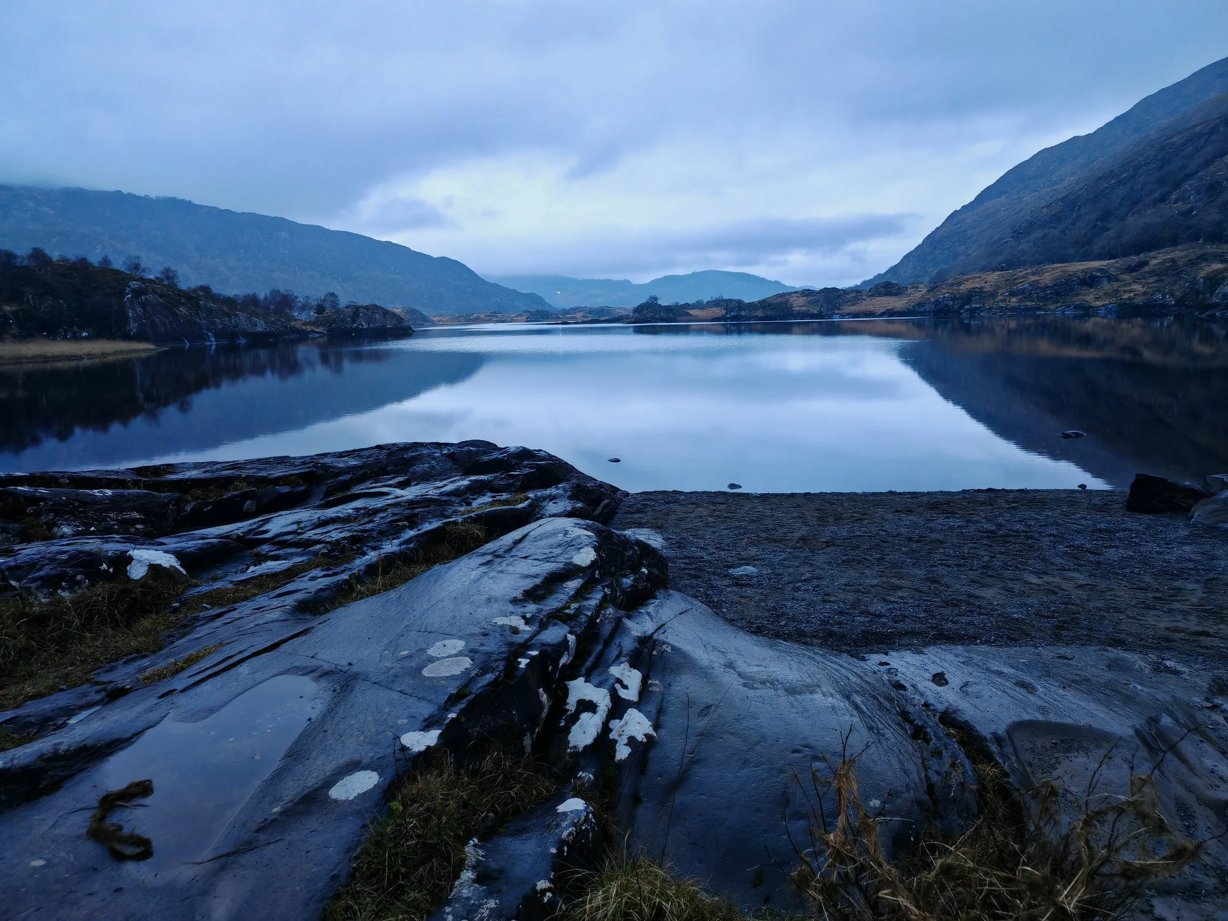 A tranquil lake surrounded by hills and mountains, with cloudy skies overhead, and a rocky shoreline in the foreground.