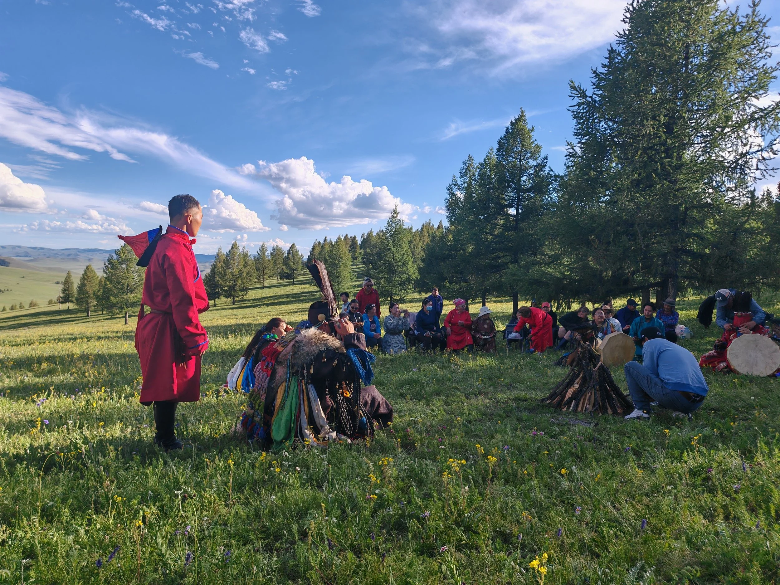 A group of people gathered outdoors on a grassy field, surrounded by trees and rolling hills under a partly cloudy sky. Some individuals are sitting, some standing, with a person in red attire prominently in the foreground.