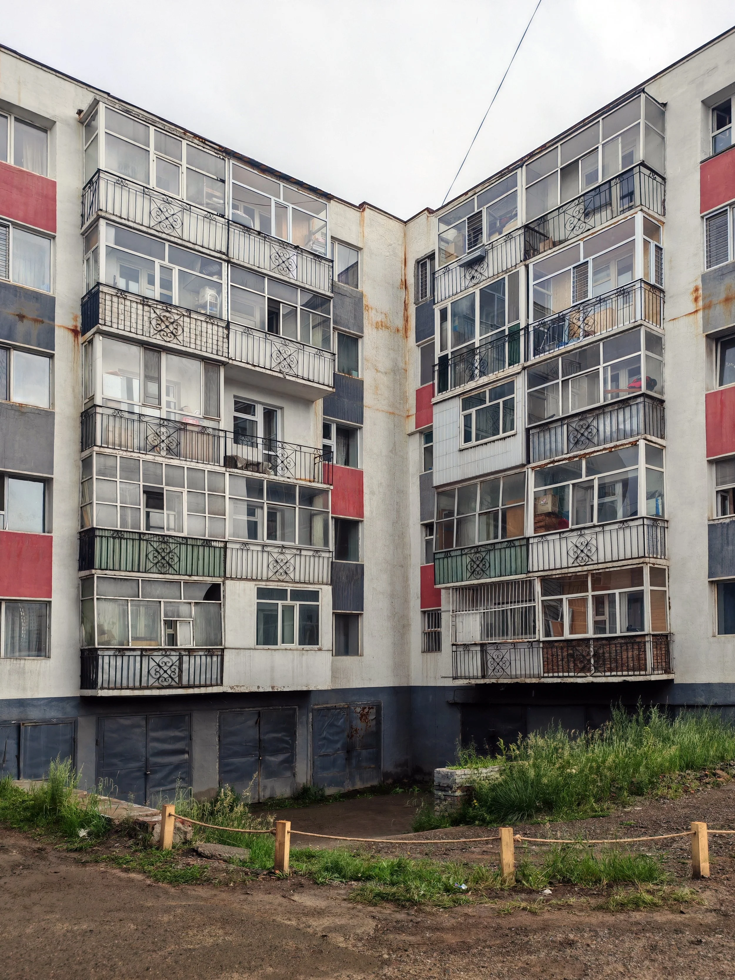 A residential apartment building with glass-enclosed balconies on each floor, showing signs of rust and wear, with a small grassy and dirt area in the foreground enclosed by a wooden fence.