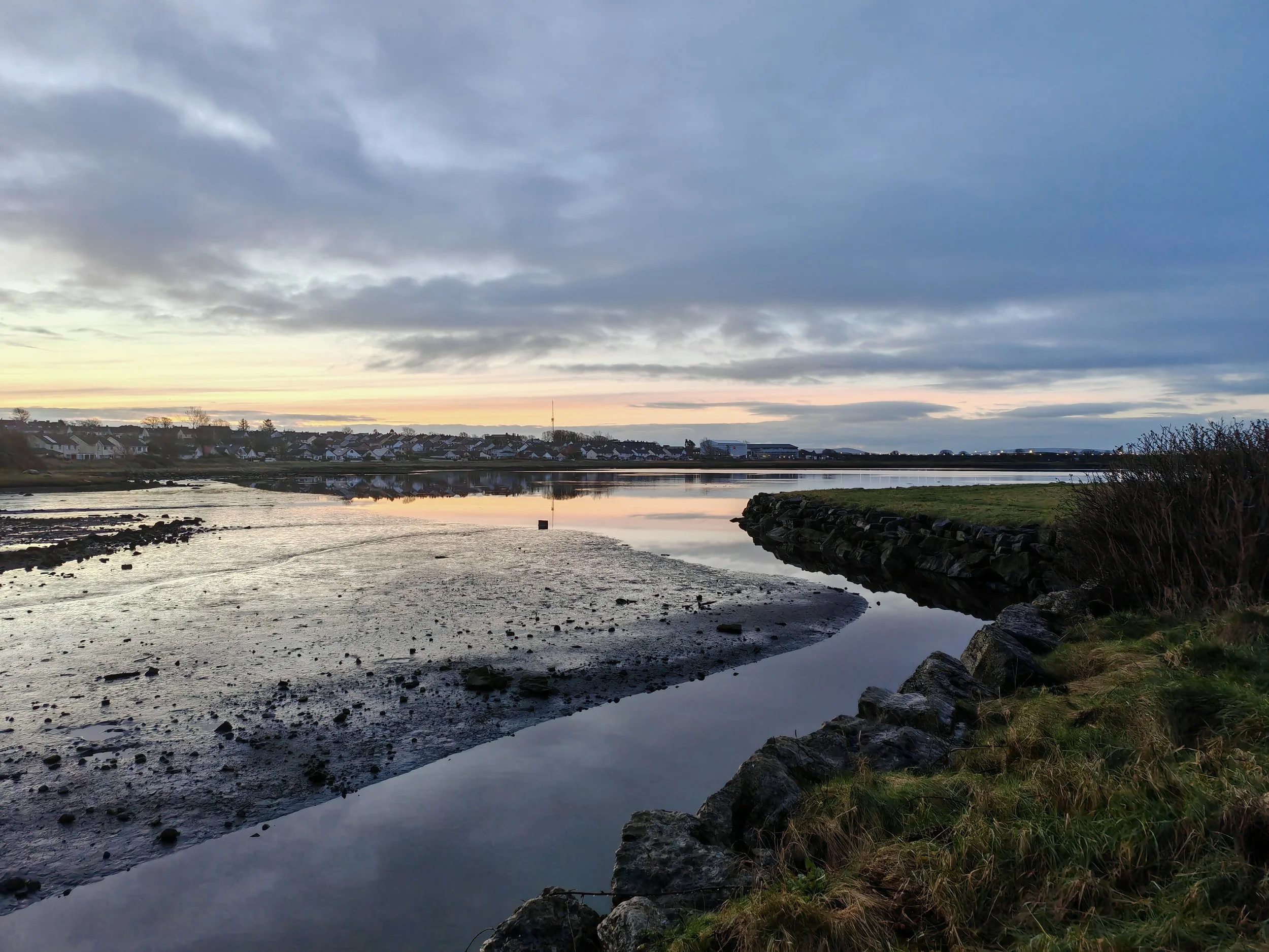 A scenic view of a river or estuary at sunset with a cloudy sky, houses on the far shore, and rocky and grassy shoreline in the foreground.