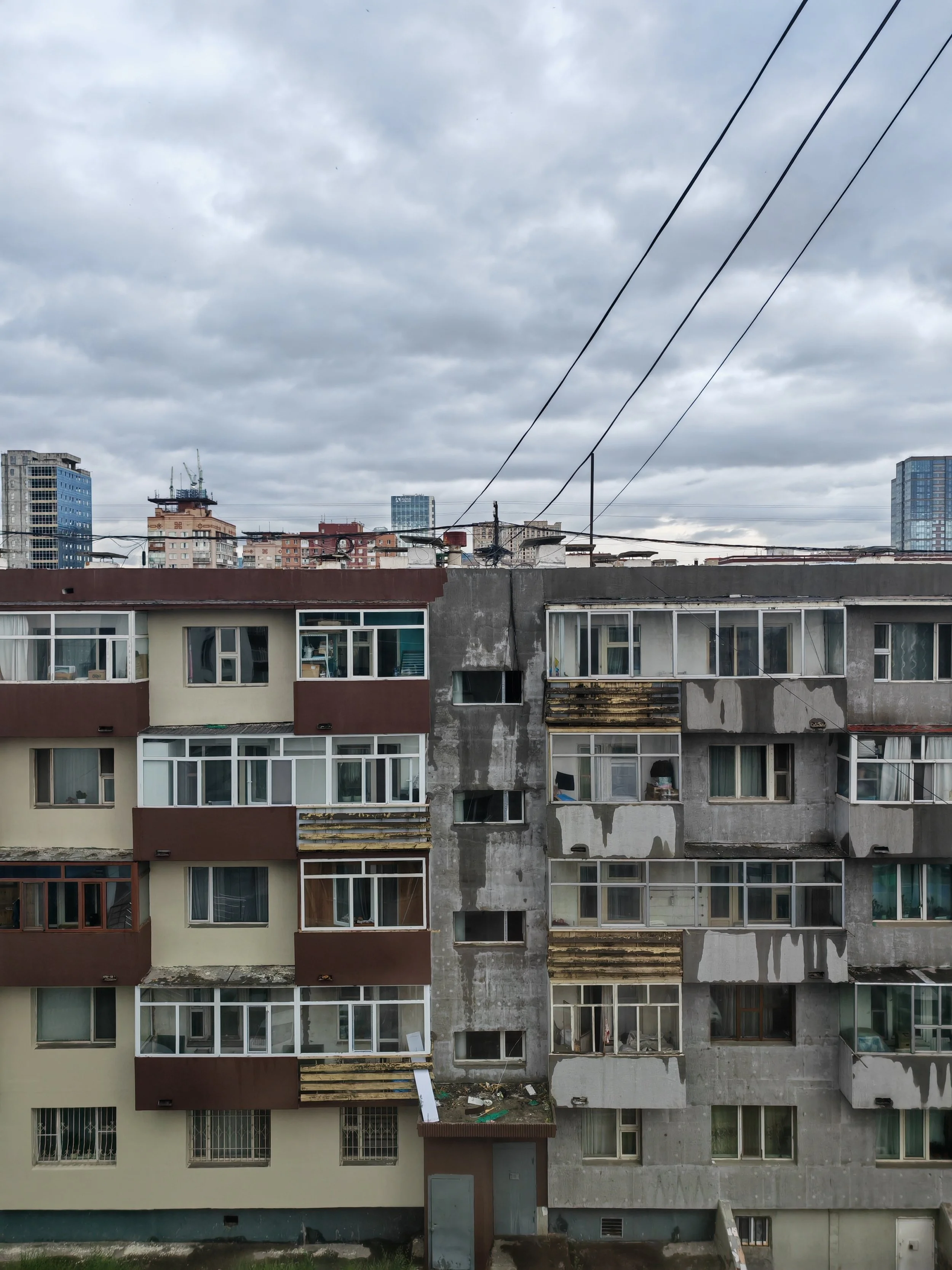 A multi-story residential building with unfinished and weathered exterior walls, windows, and balconies, with prominent overhead power lines against a cloudy sky.