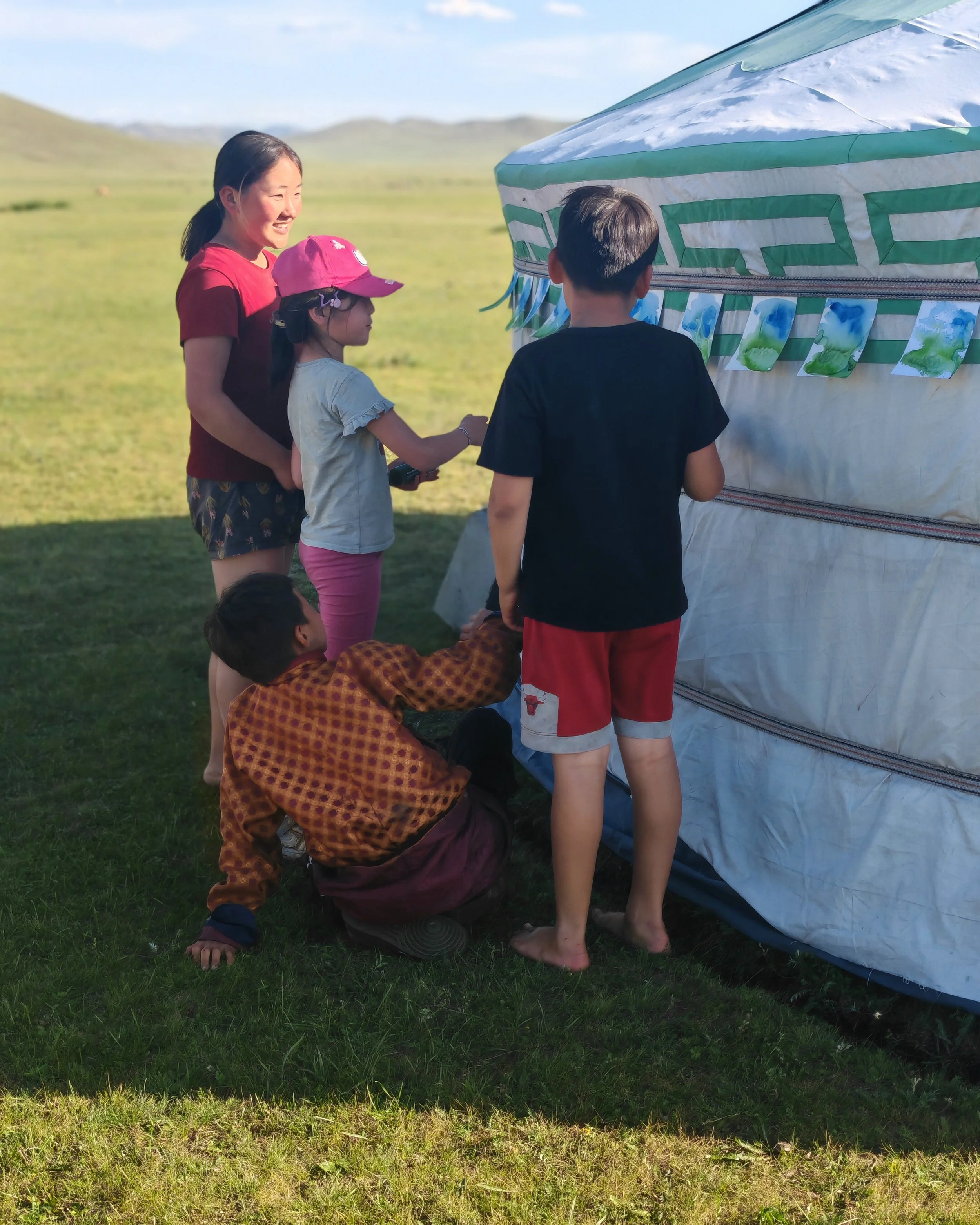 Four children, two girls and two boys, gathered around a colorful, patterned tent outdoors on a grassy field. The girl in a red shirt and another girl with a pink cap are looking at photographs posted on the tent, while a boy in a black shirt and red