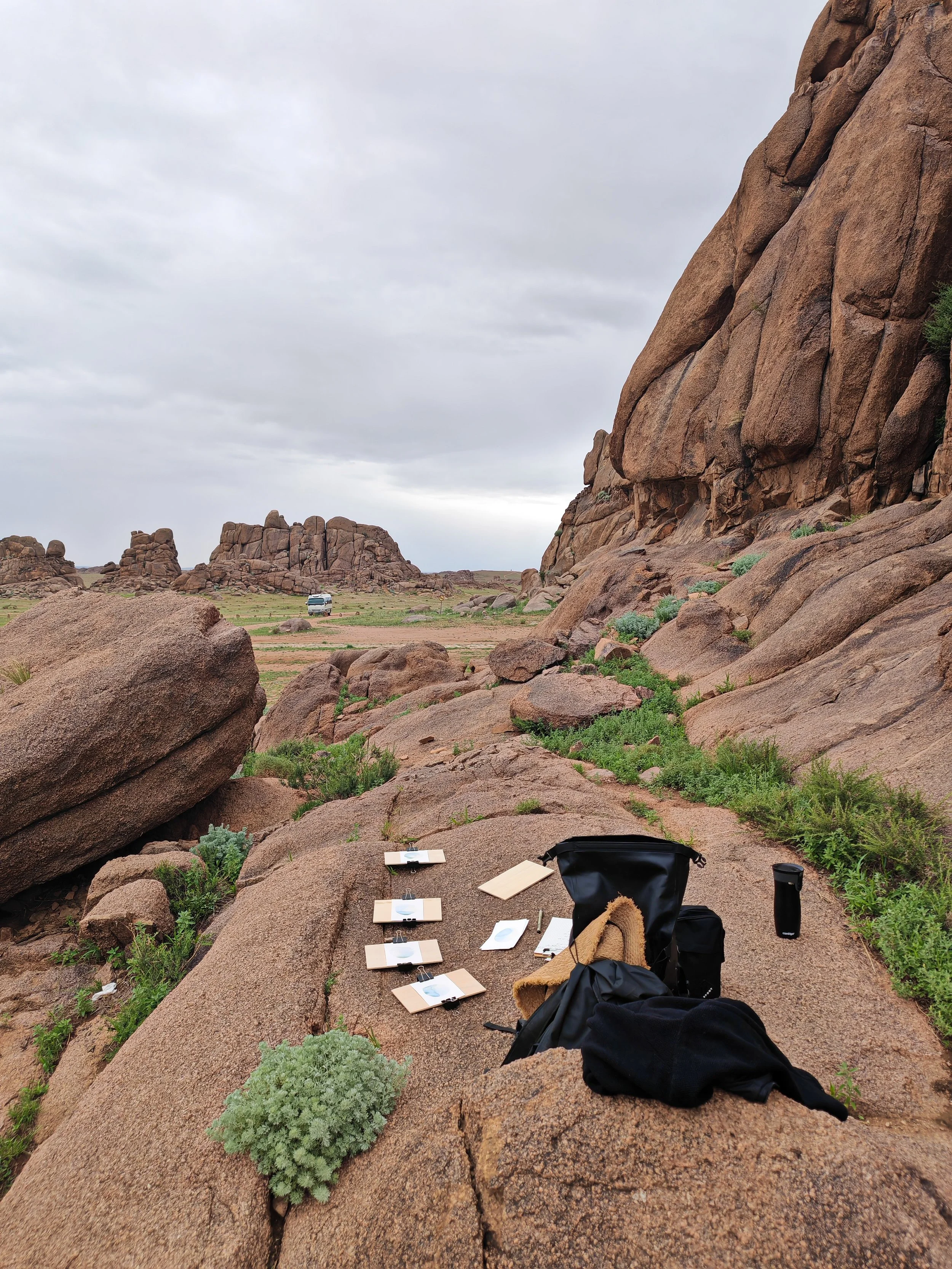 Climbing gear and papers laid out on a large rocky surface in a desert landscape with large rock formations and cloudy sky.