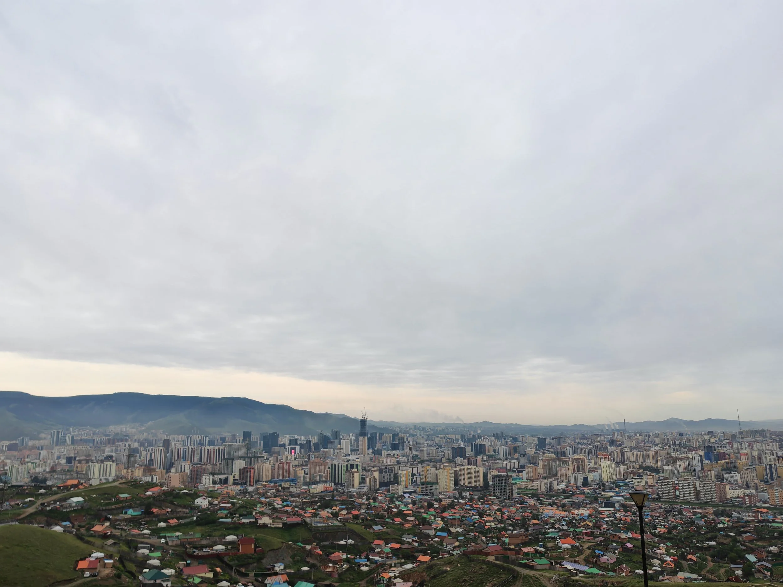 Cityscape with high-rise buildings and surrounding residential houses, with mountains in the background and cloudy sky overhead.