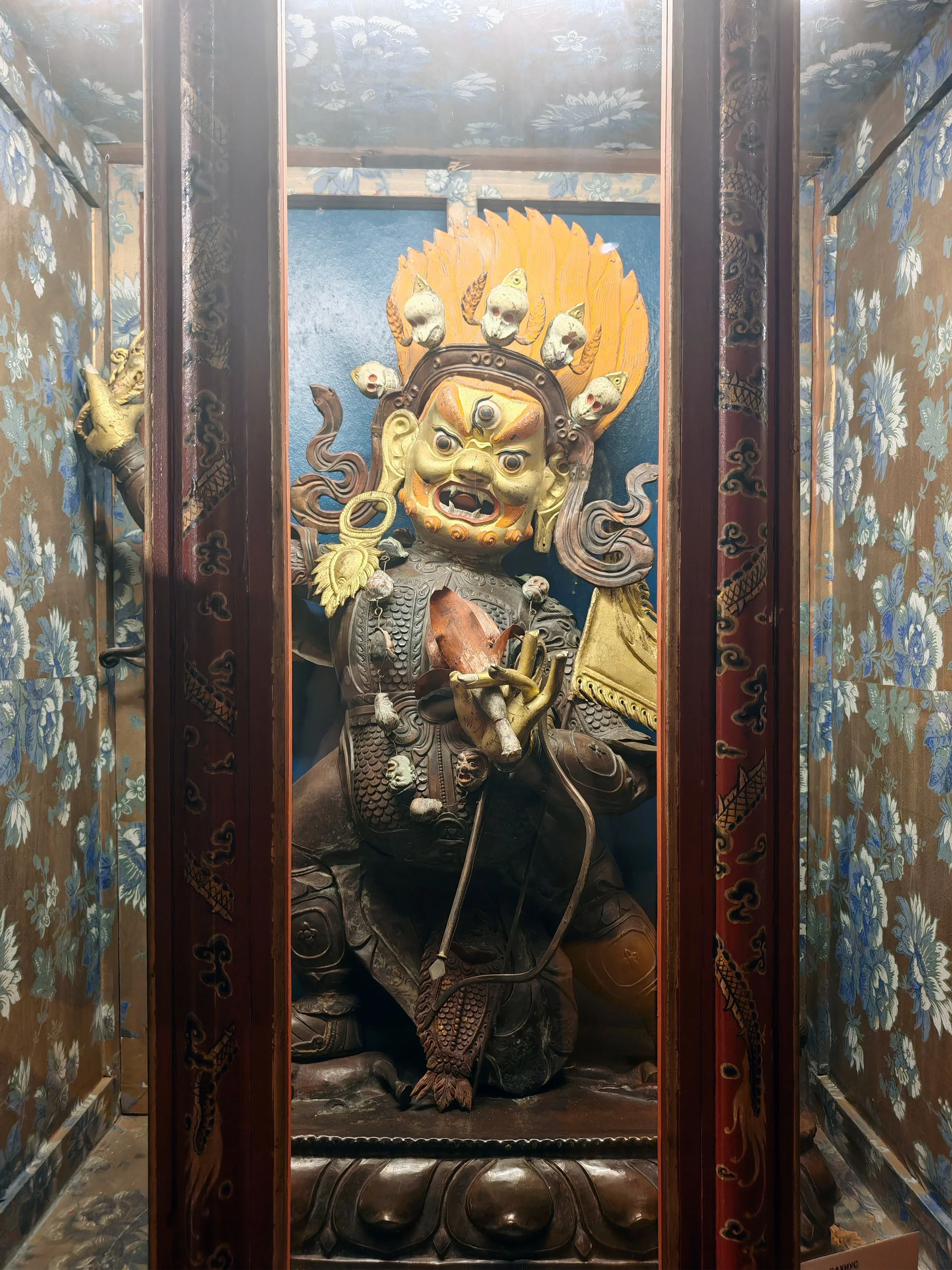 A detailed statue of a fierce mythological warrior or deity with multiple skulls on his headdress, holding a weapon, and surrounded by ornate decorations, placed inside a shrine with floral patterned walls and wooden framing.