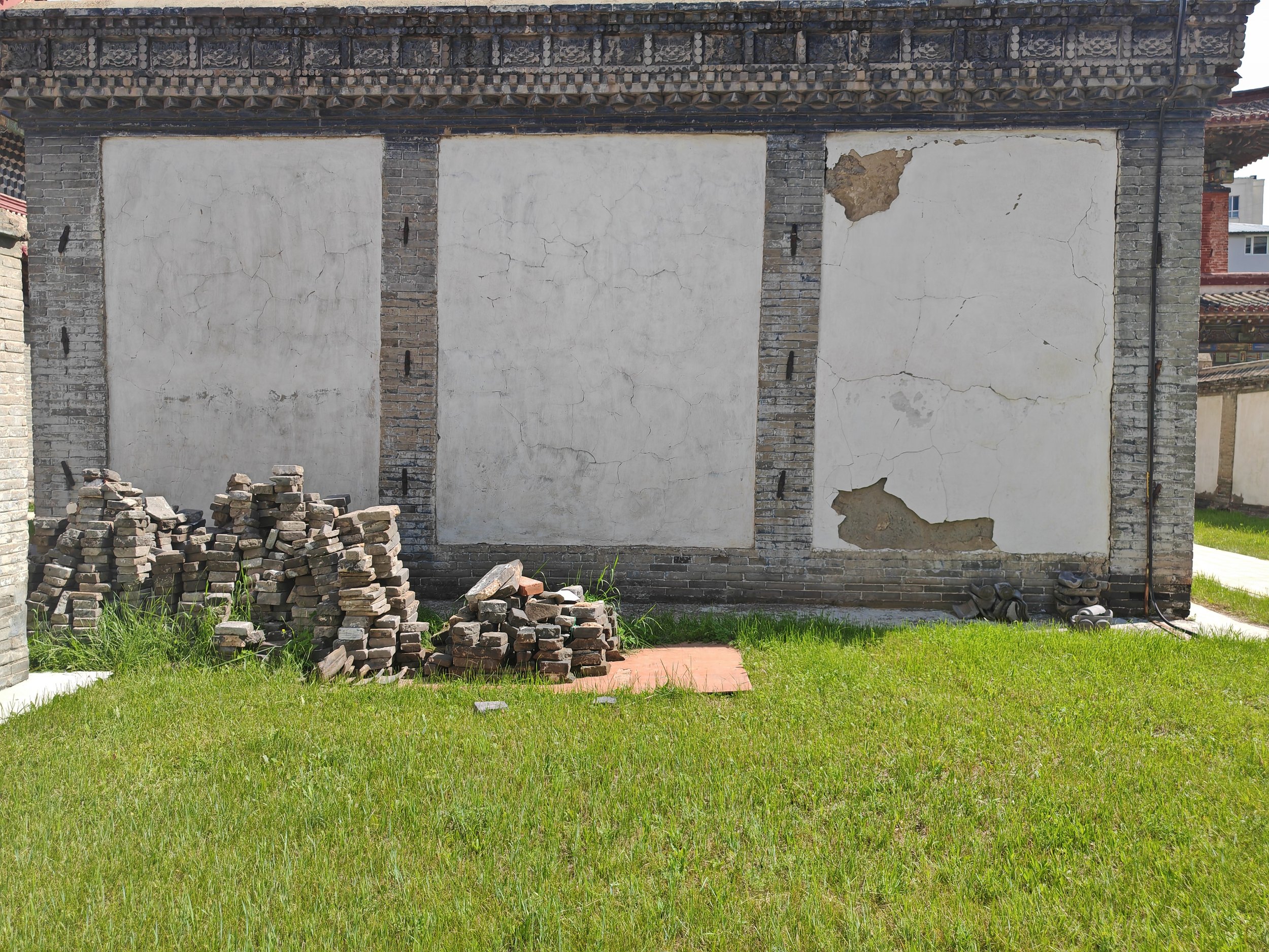 A house with a white wall and black wooden beams, showing cracks and damage, with a pile of bricks and grass in front.