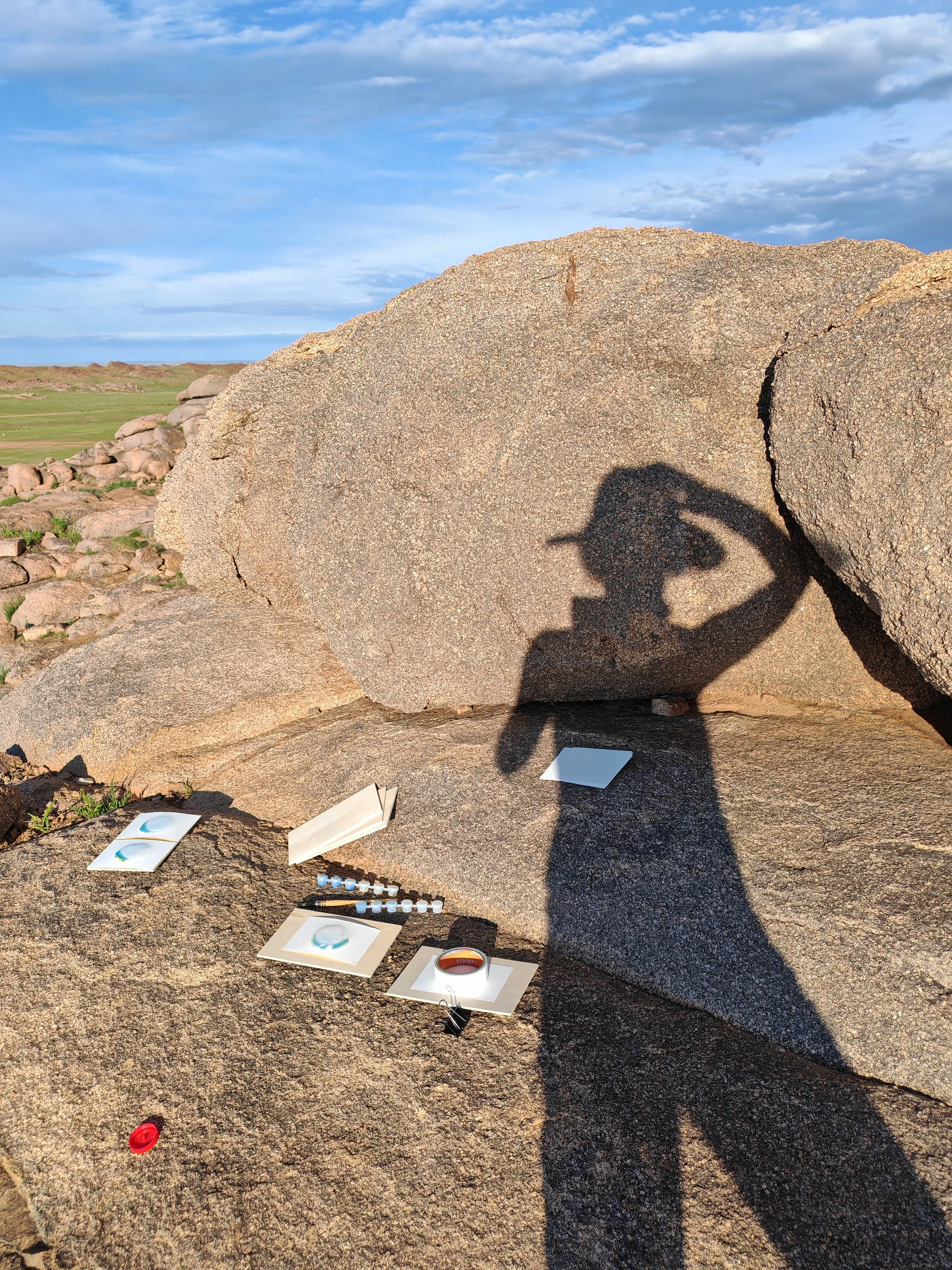 Shadow of a person taking a photo on a rocky landscape with scattered papers and objects on the ground, under a partly cloudy sky.