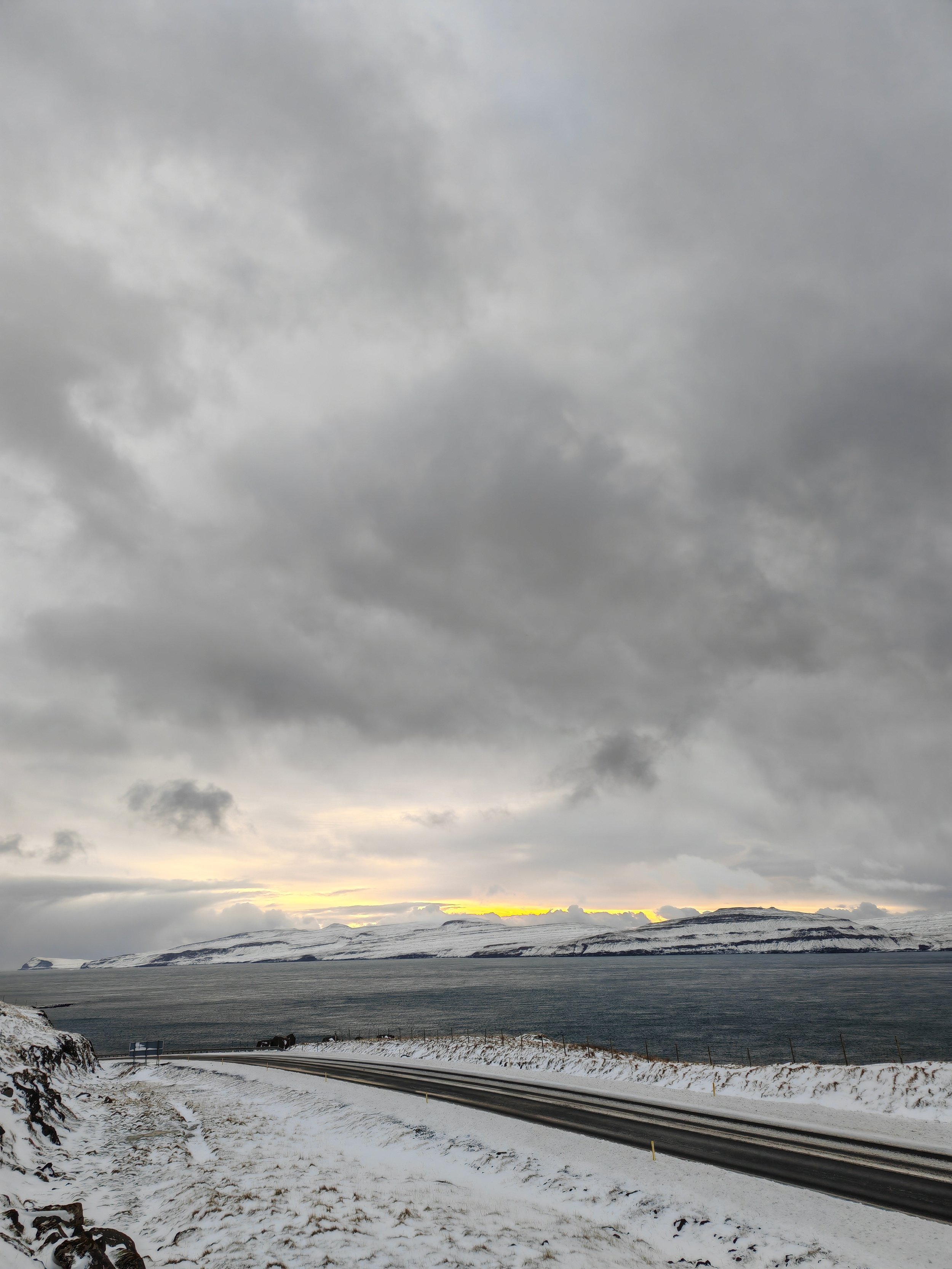 A snowy landscape with a winding road, body of water, and snow-covered hills or mountains under a cloudy sky at sunset or sunrise.