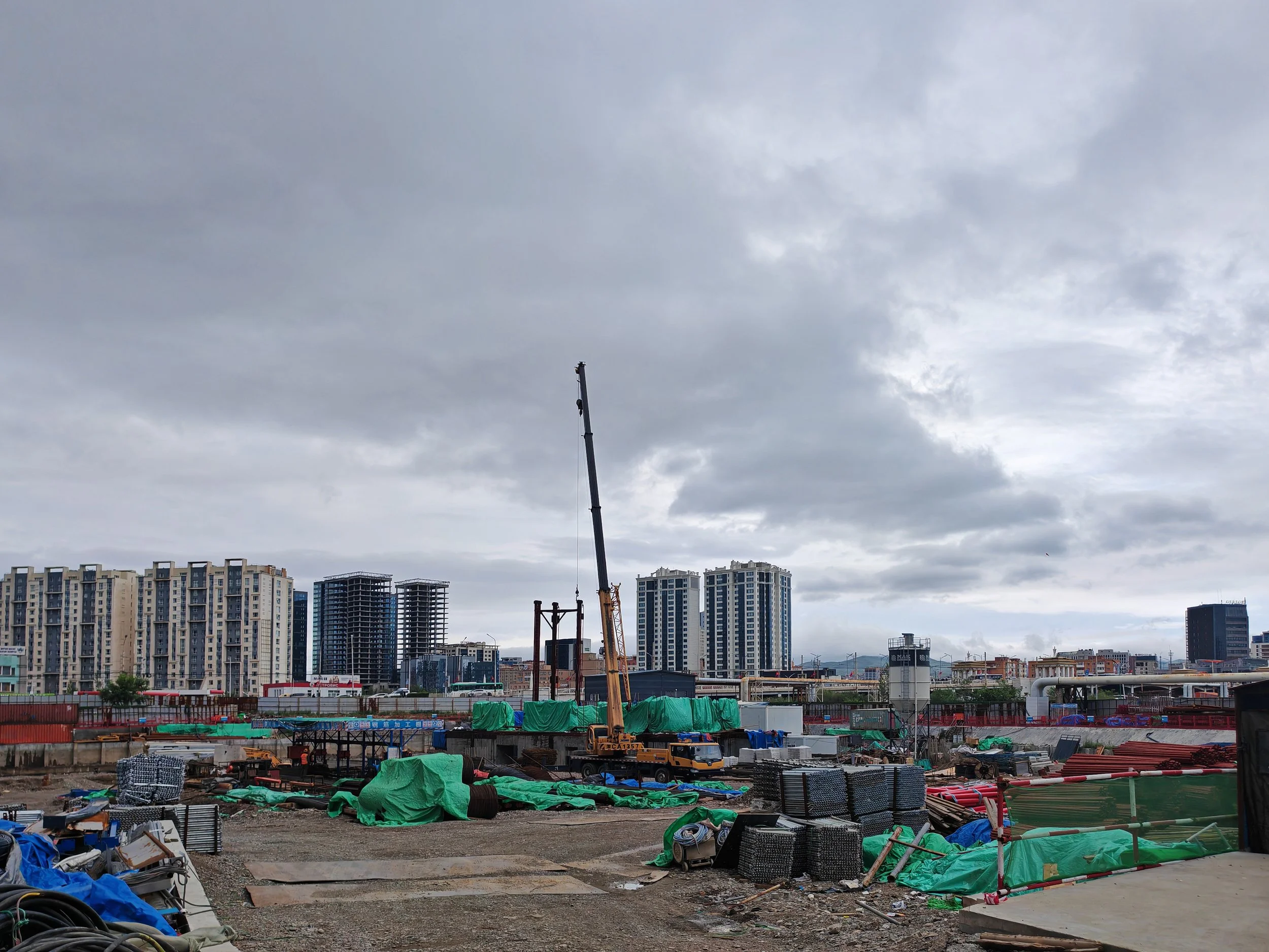 A construction site with a crane lifting a large object, surrounded by unfinished buildings and construction materials covered with green tarps, with a backdrop of completed high-rise residential buildings under a cloudy sky.