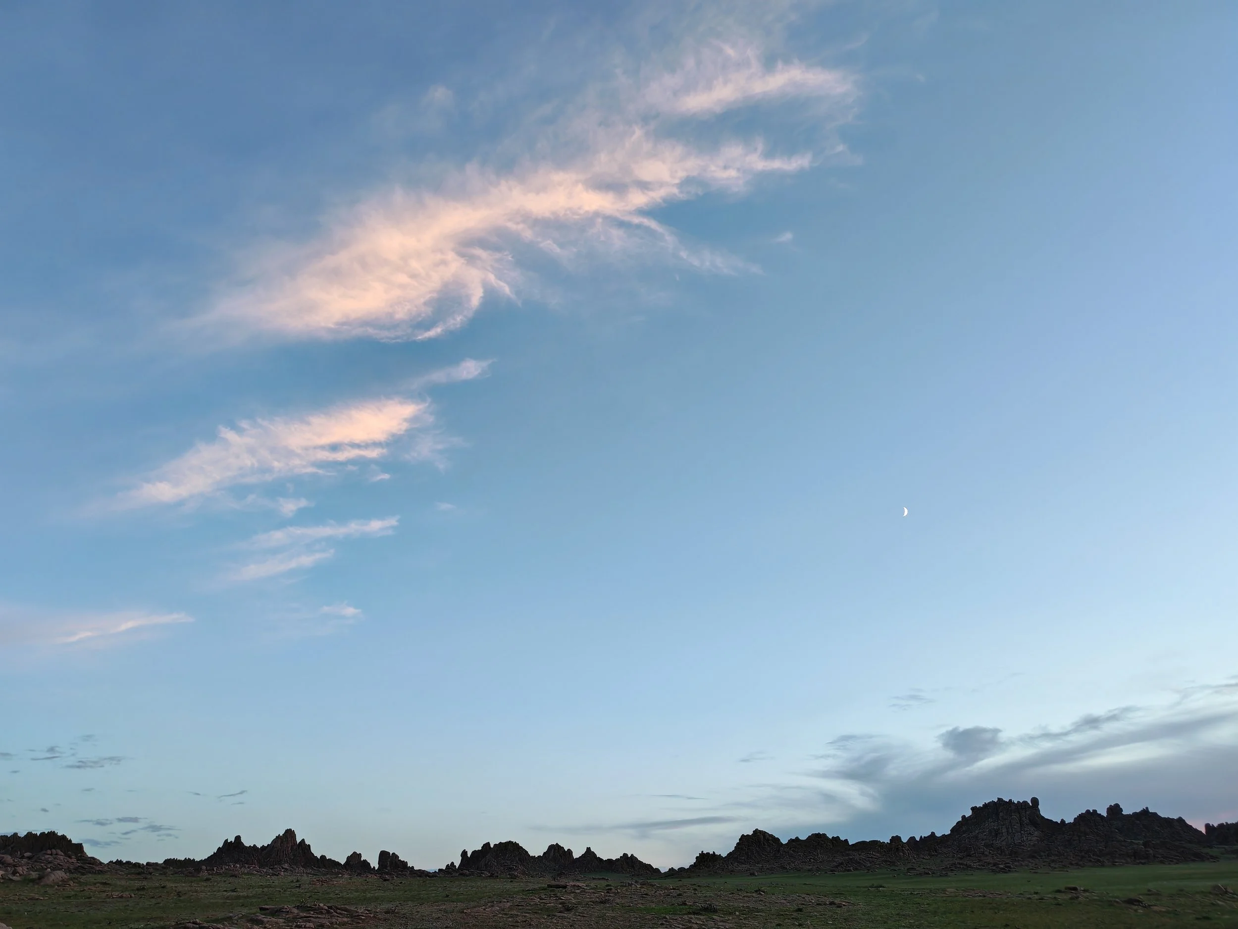 A wide view of a landscape with rugged mountains, a grassy plain, and a blue sky with wispy clouds and a visible crescent moon.