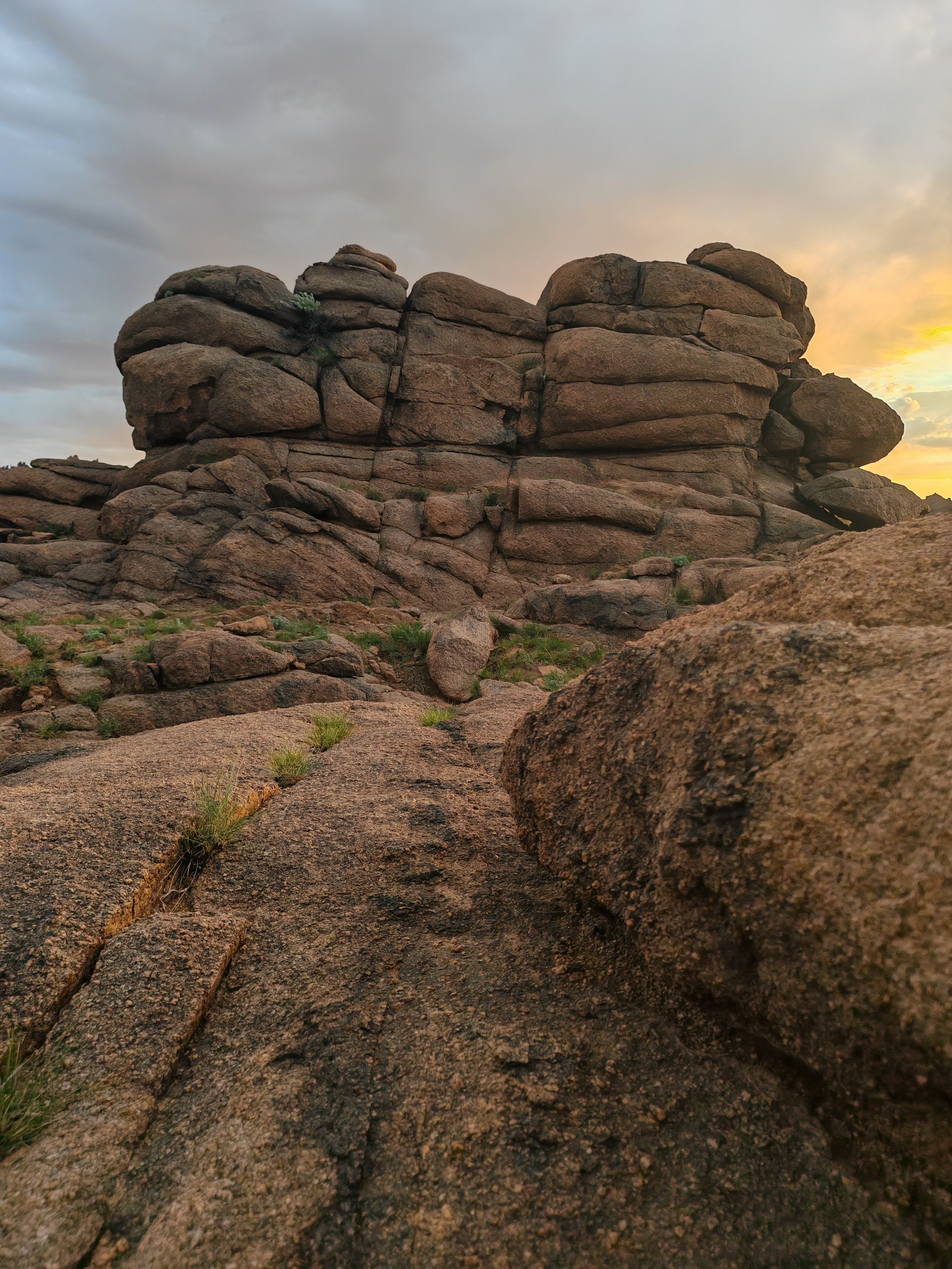 A large formation of rocks and boulders in a desert landscape during sunset or sunrise, with a cloudy sky overhead.