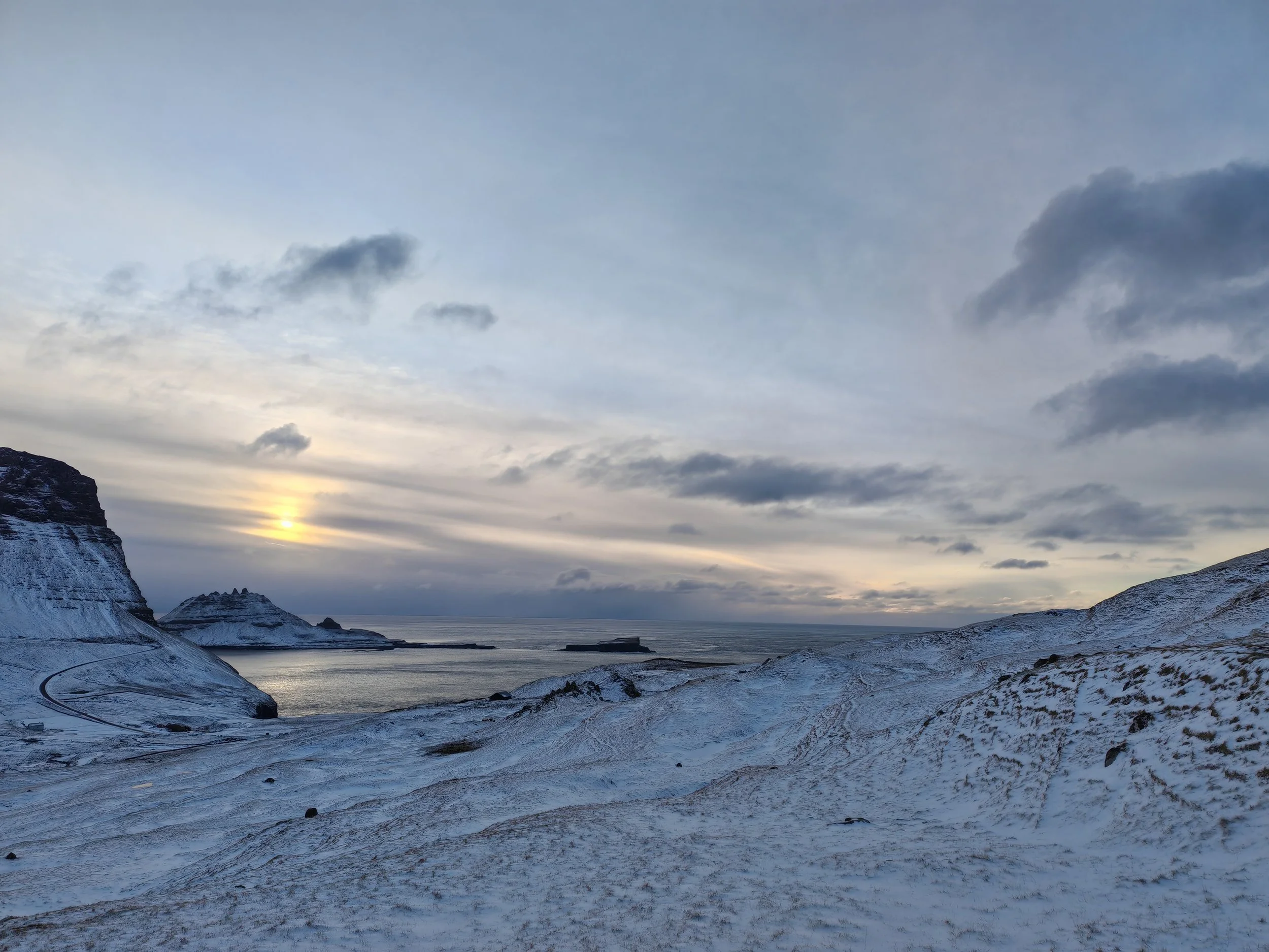 A snowy landscape with hills sloping down toward a body of water, under a cloudy sky with a setting or rising sun on the horizon.