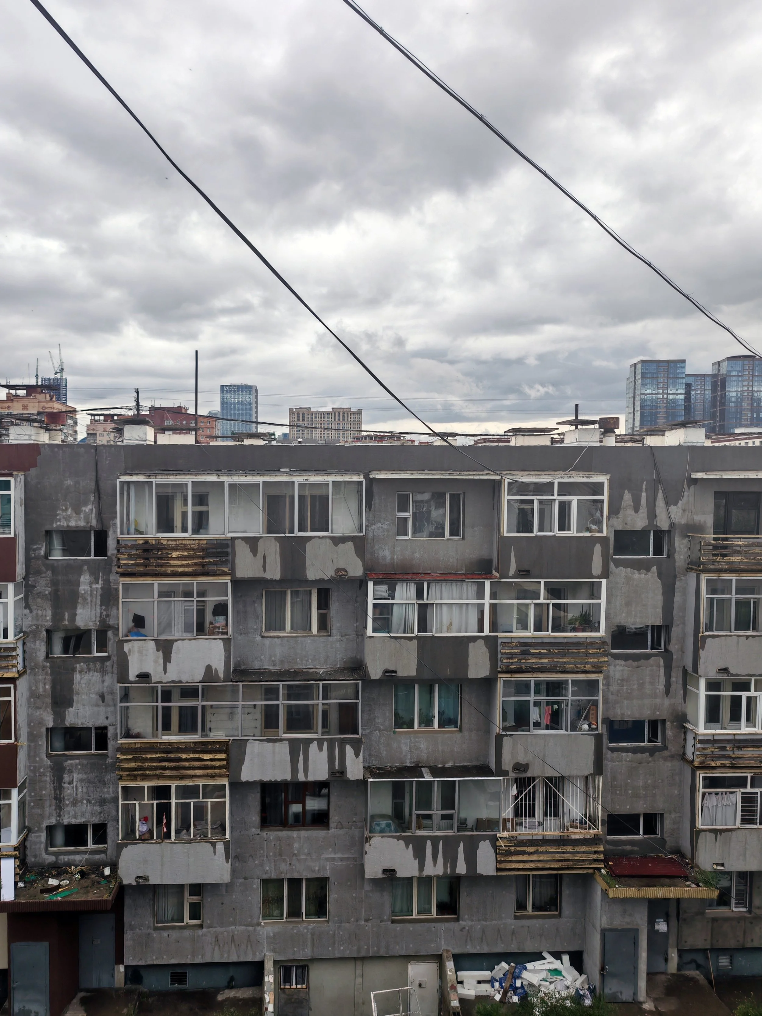 A multi-story apartment building with a gray, weathered exterior, laundry hanging inside the windows, and a small yard with discarded construction materials at the bottom.