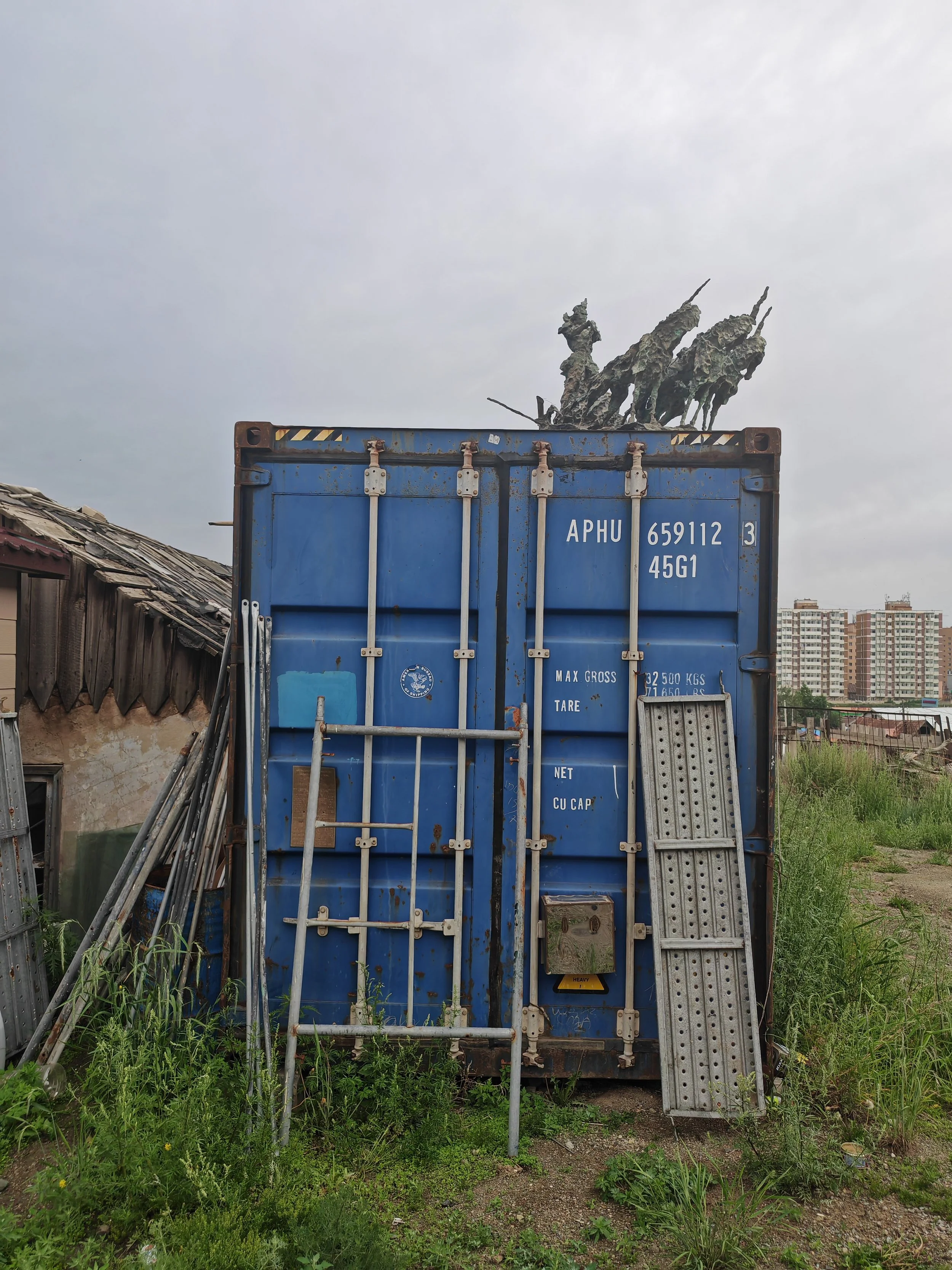 An open blue shipping container with rust and dirt, topped with three large, weathered statues of soldiers on horseback, in a grassy area near abandoned structures and high-rise buildings in the background.