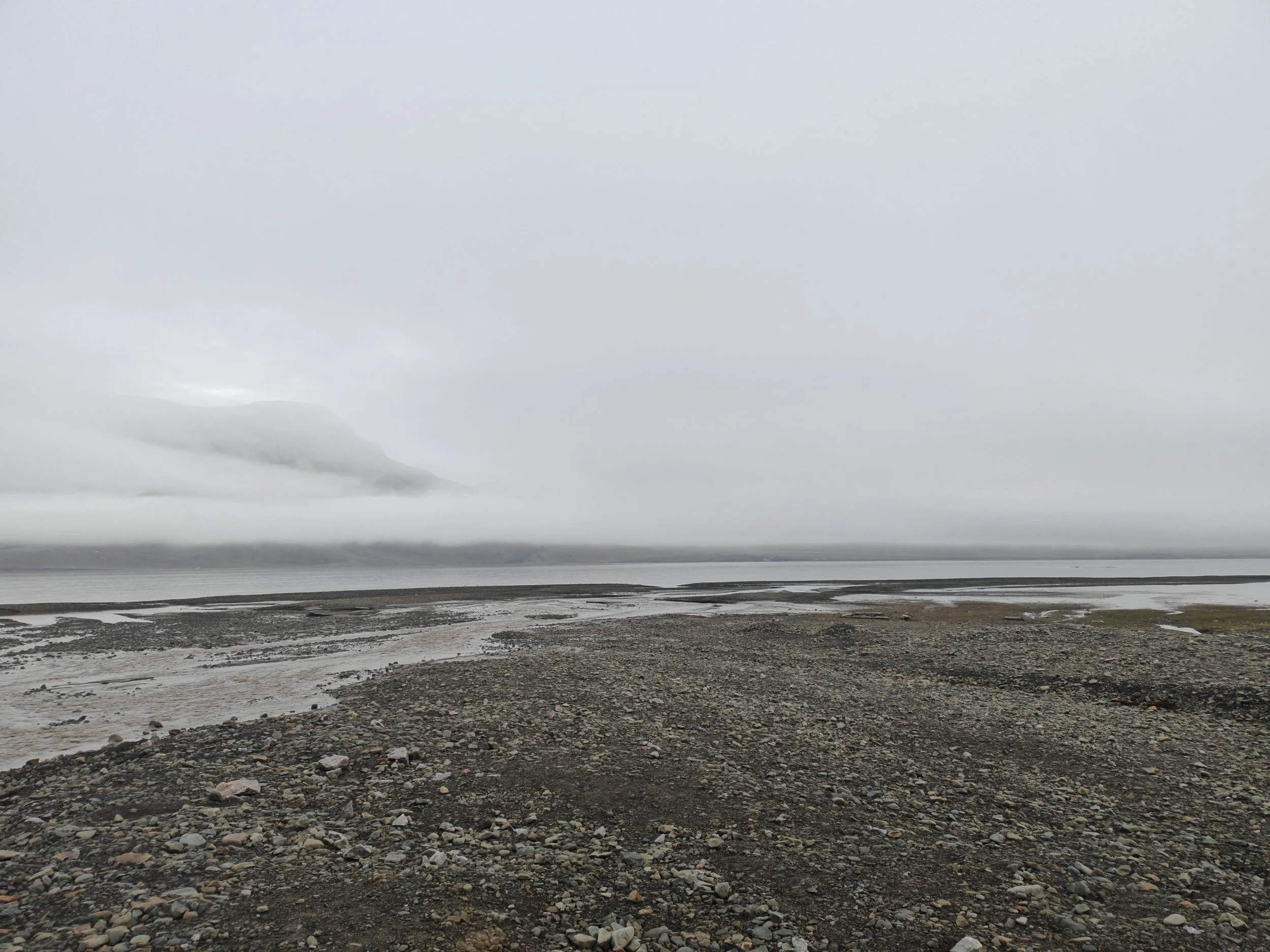 A vast, overcast landscape with a rocky shoreline in the foreground and a calm body of water extending to the horizon. Mountains are faintly visible in the distance under a cloudy sky.
