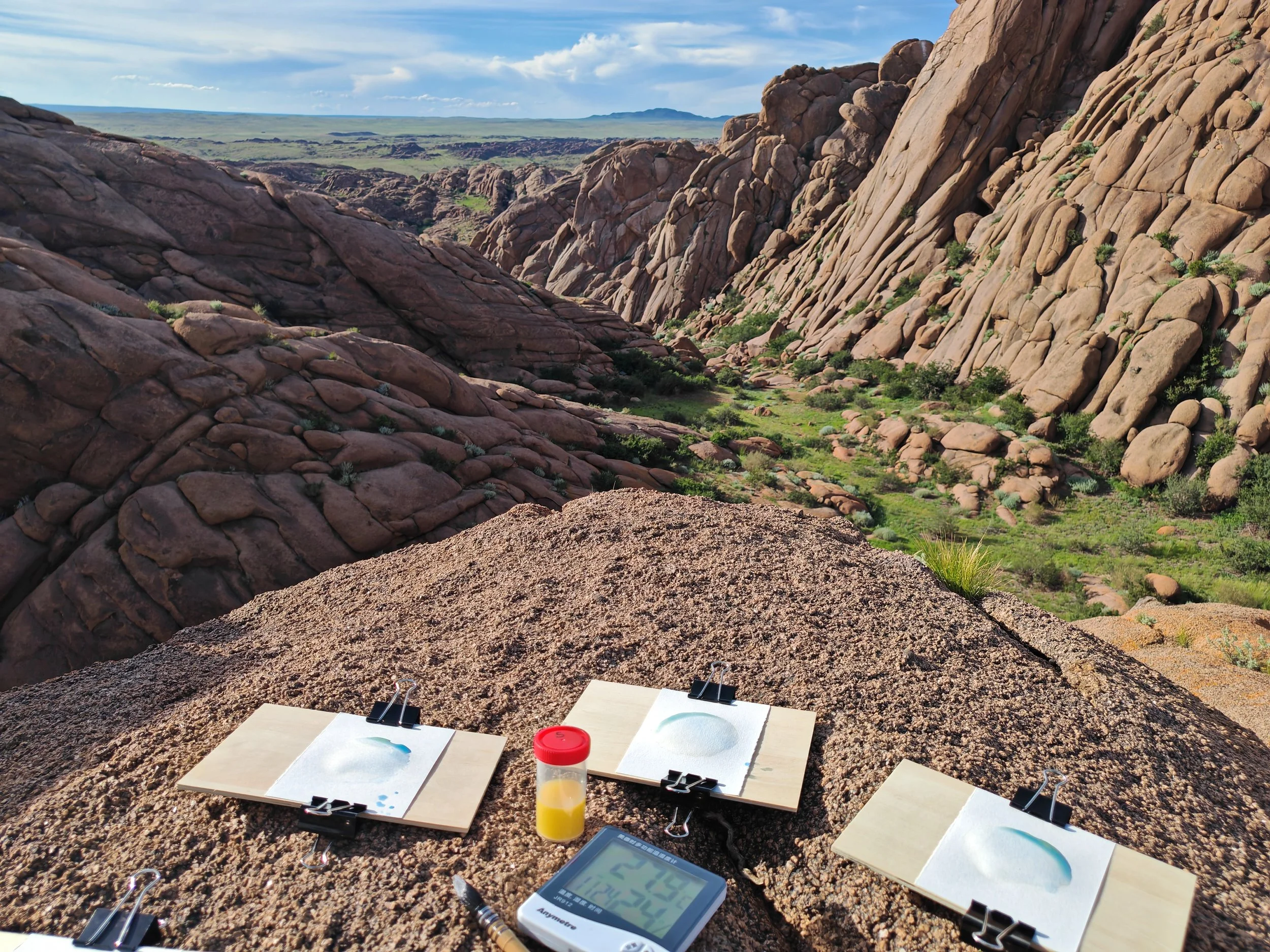 Cliffs and rocky landscape under a blue sky with some clouds, with scientific equipment on a flat rock in the foreground.