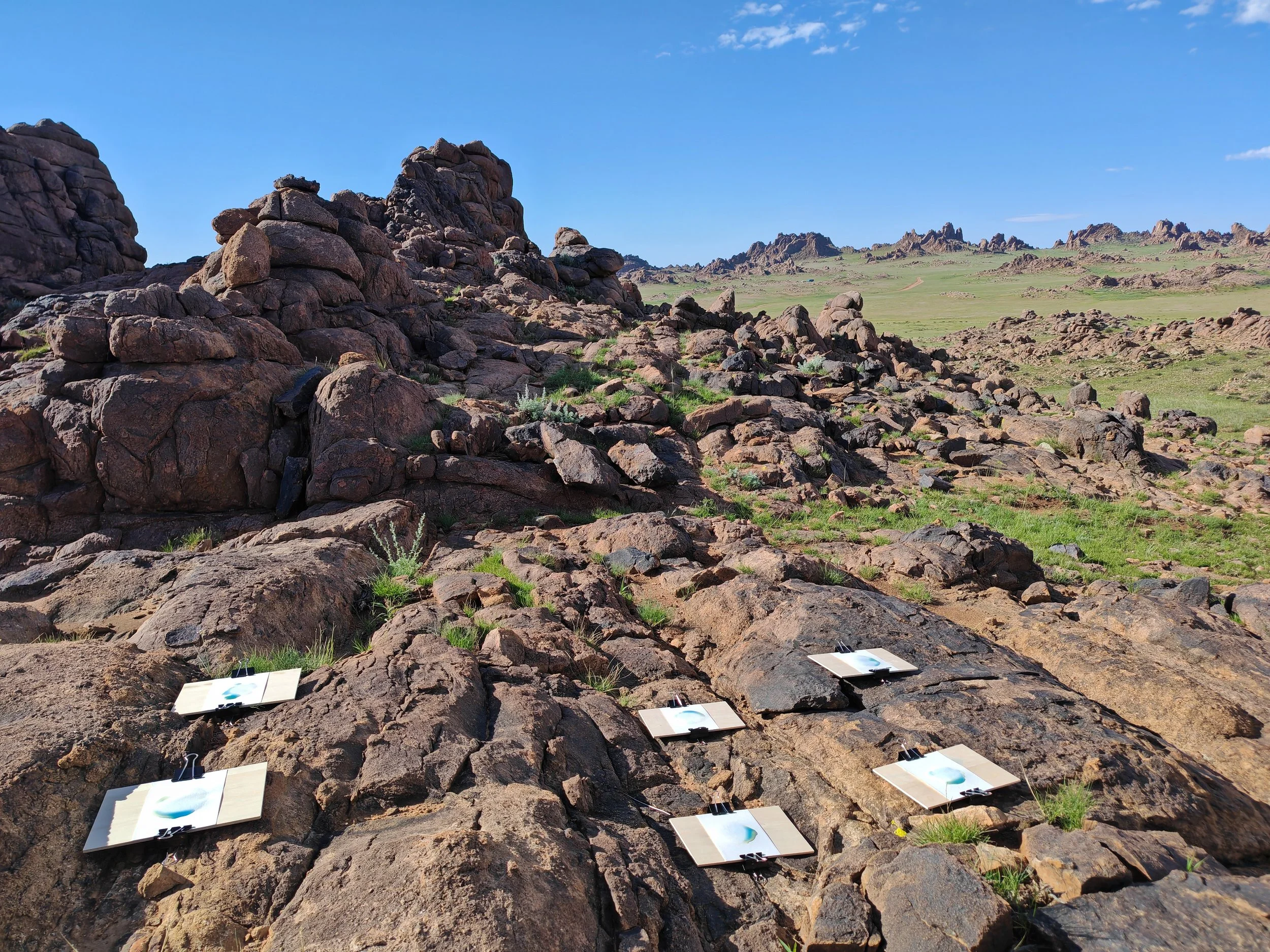A desert landscape with rocky formations and green grass, under a clear blue sky, with several white cards attached to the rocks with black clips.