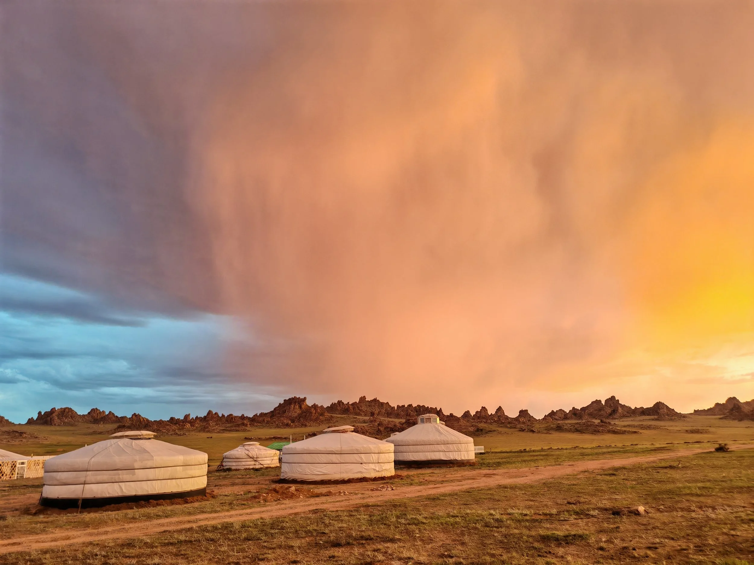 A landscape of yurts on grassy plain with rocky hills and colorful sky at sunset.