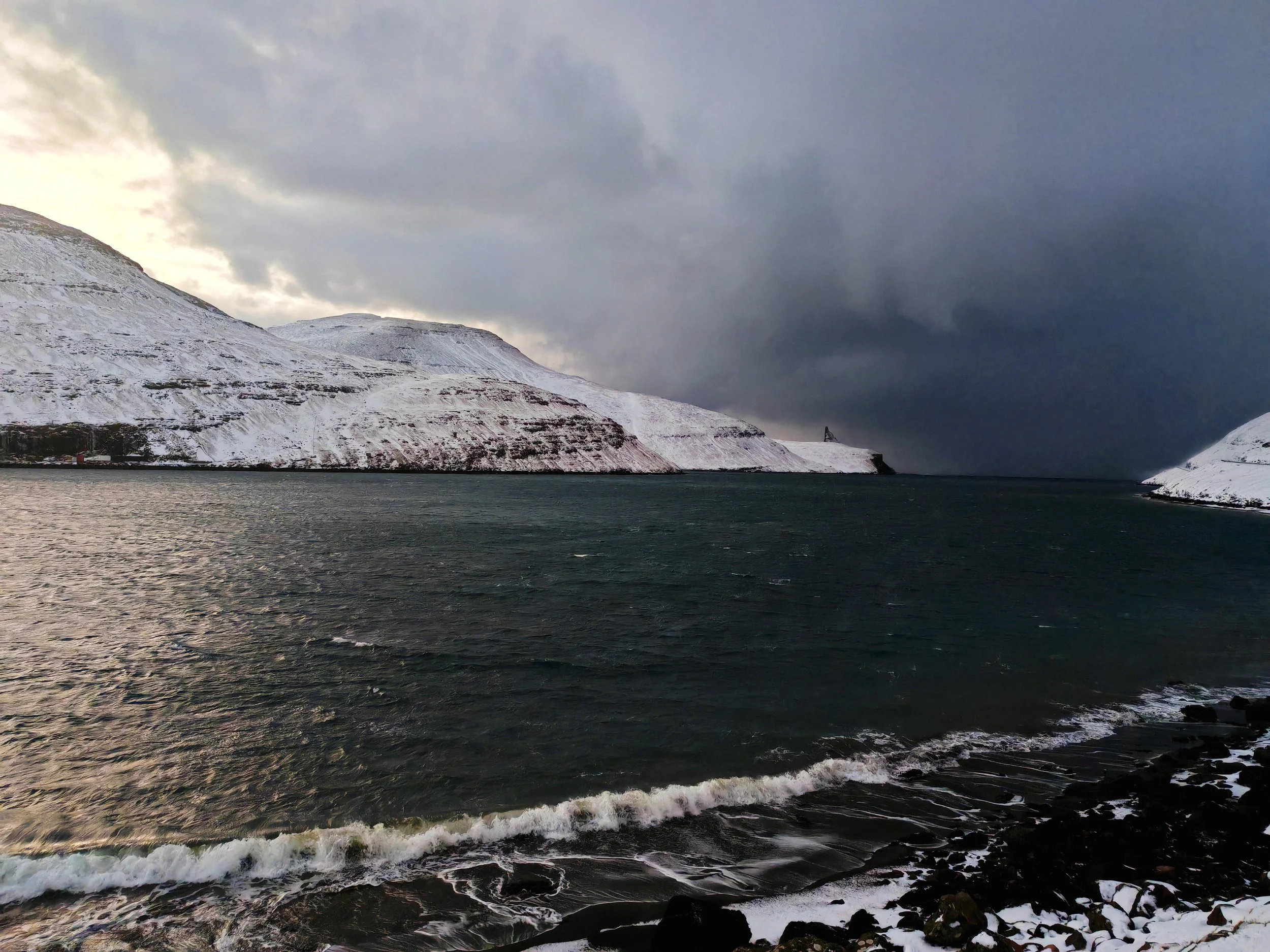 Snow-covered mountains beside a dark body of water under a cloudy sky.