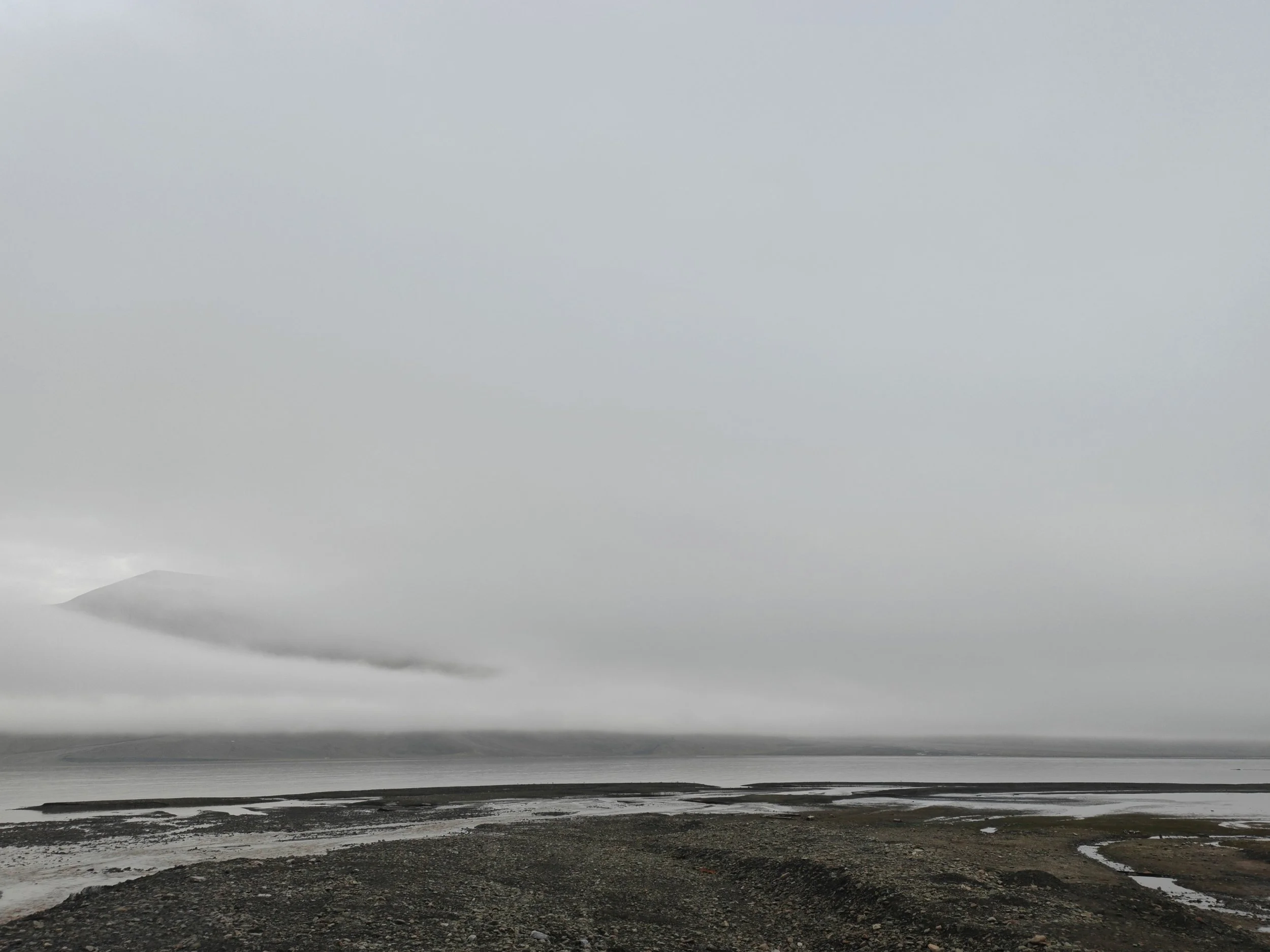 A desolate, cloudy beach with dark wet stones and patches of sand, and a large body of water extending to the horizon under a gray sky.