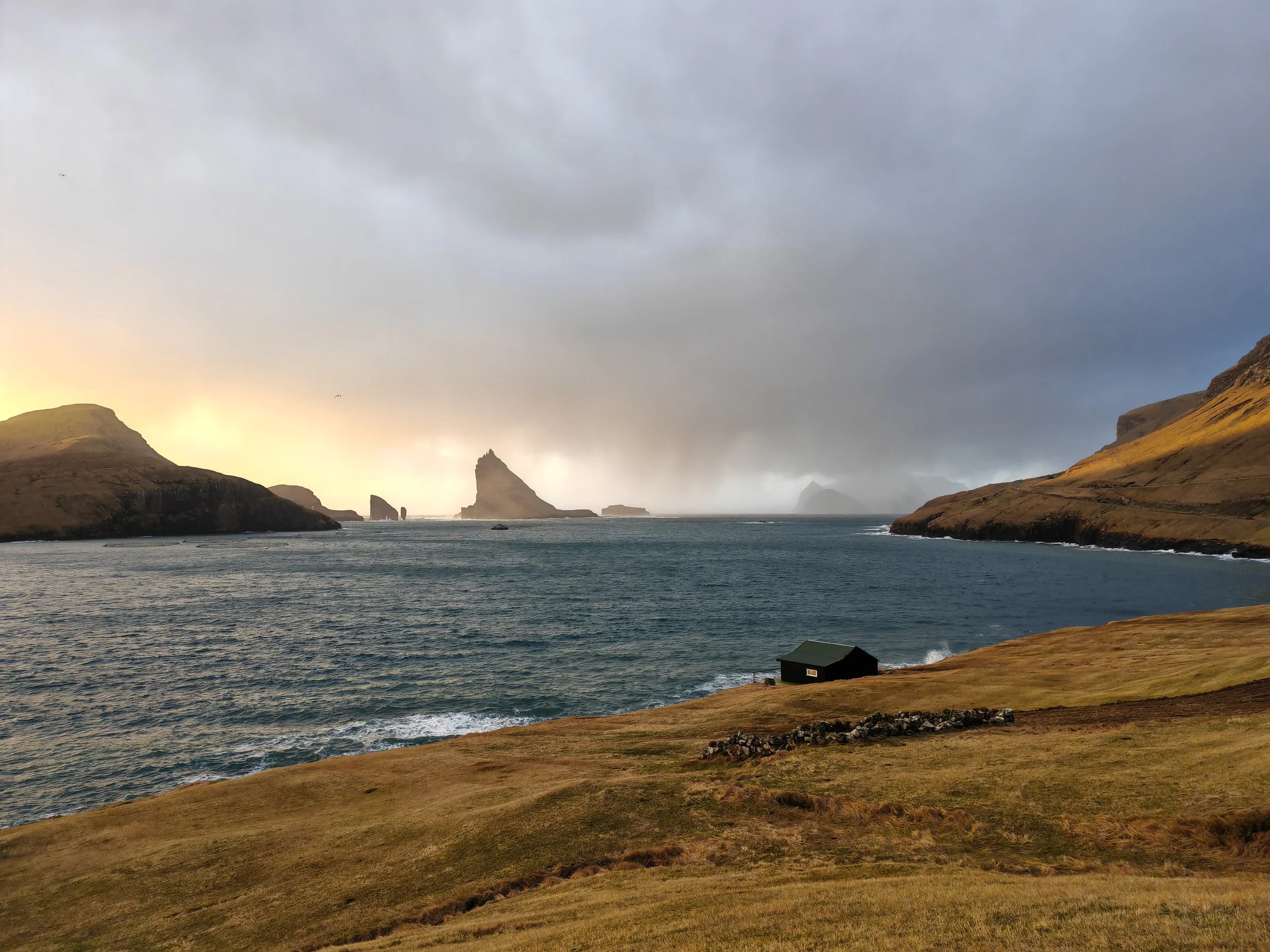 A coastal landscape with rolling grassy hills leading to a body of water. There are rocky formations or small islands in the distance with a cloudy sky overhead.