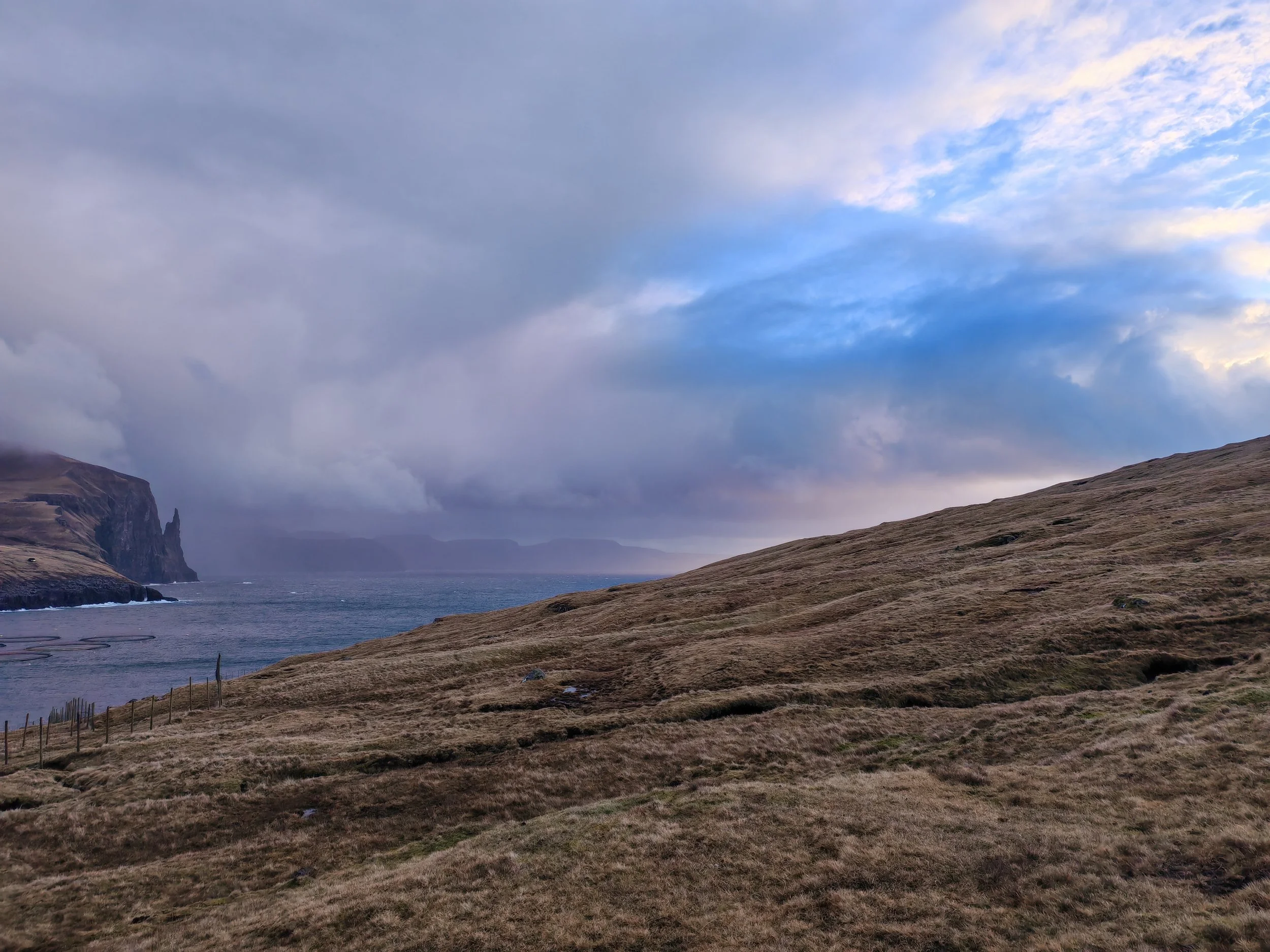 A coastal landscape with grassy hills, a rocky cliff in the distance, and a body of water under a cloudy sky with patches of blue.