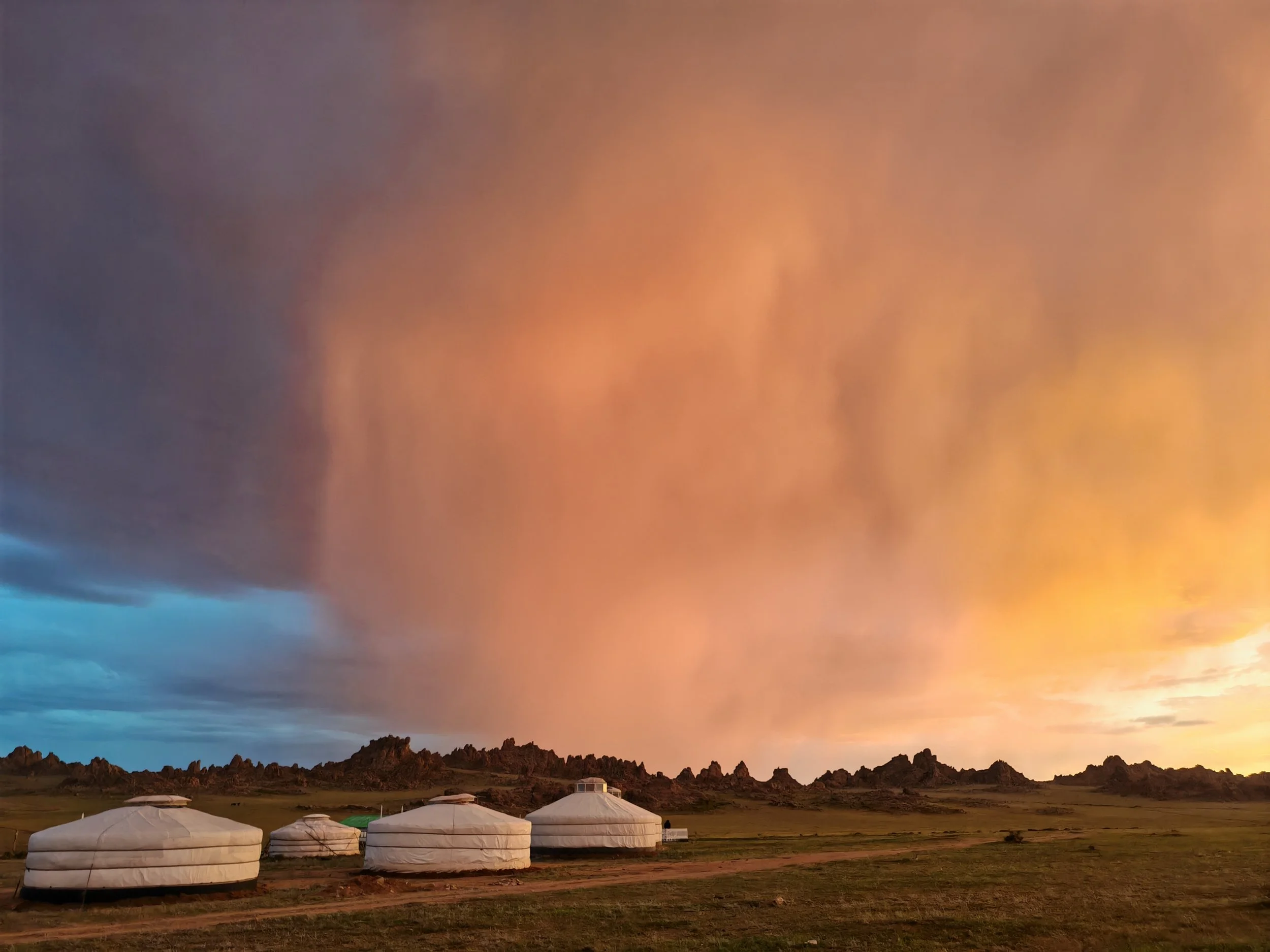 A landscape with yurts in the foreground, rocky hills or mountains in the background, and a colorful sunset sky with clouds.