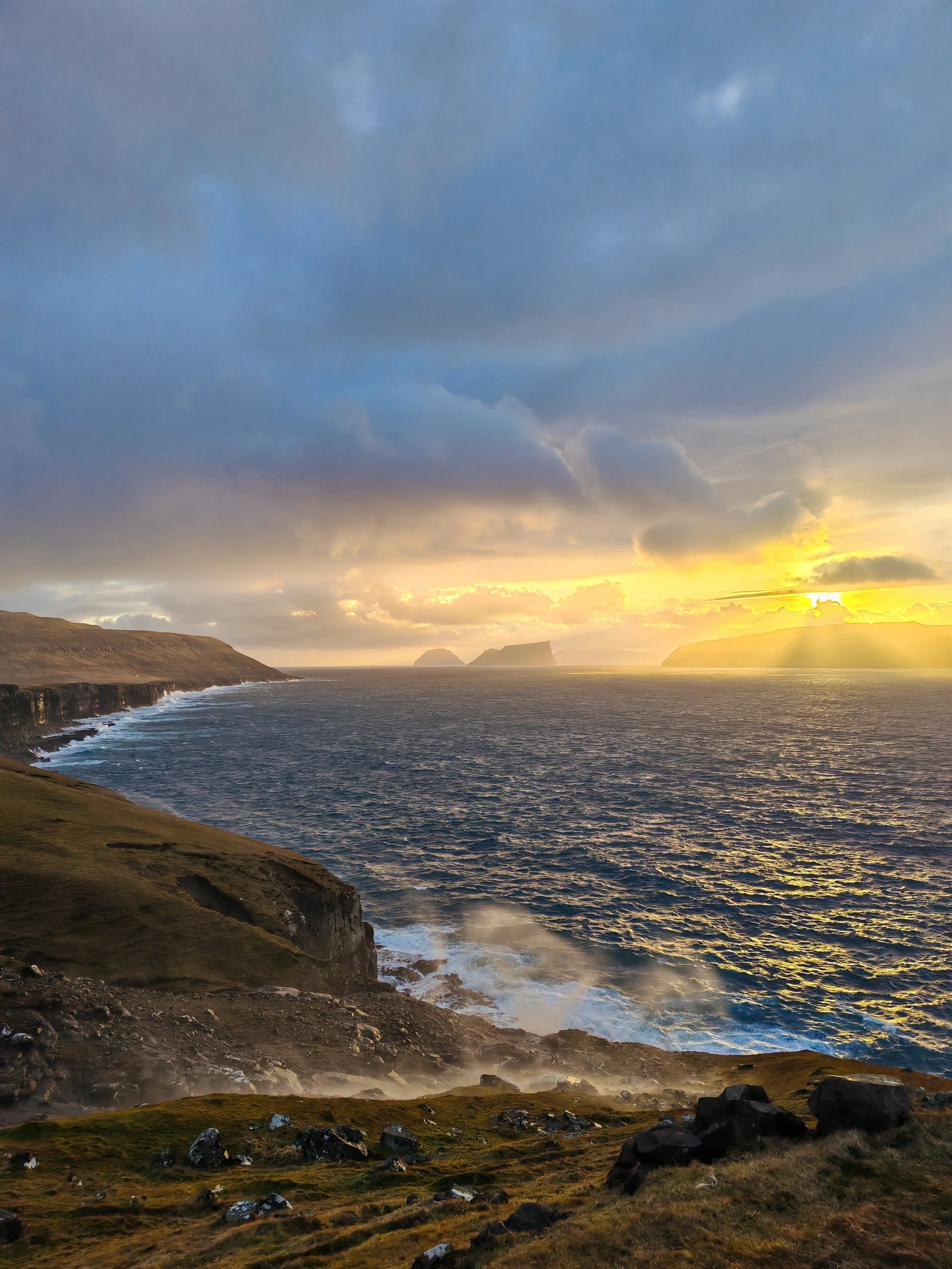 Cliffside overlooking ocean with cloudy sky and setting sun, distant islands visible across water.