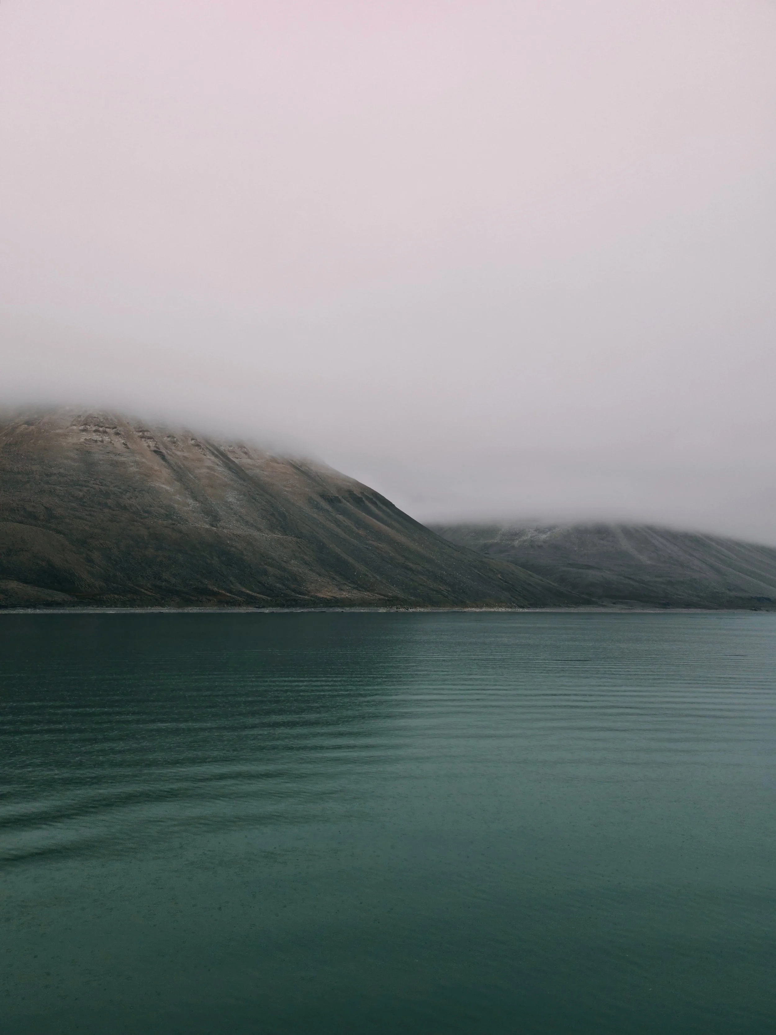 Scenic view of a calm body of water with mountains in the background, partially covered by low-hanging clouds and mist, under an overcast sky.