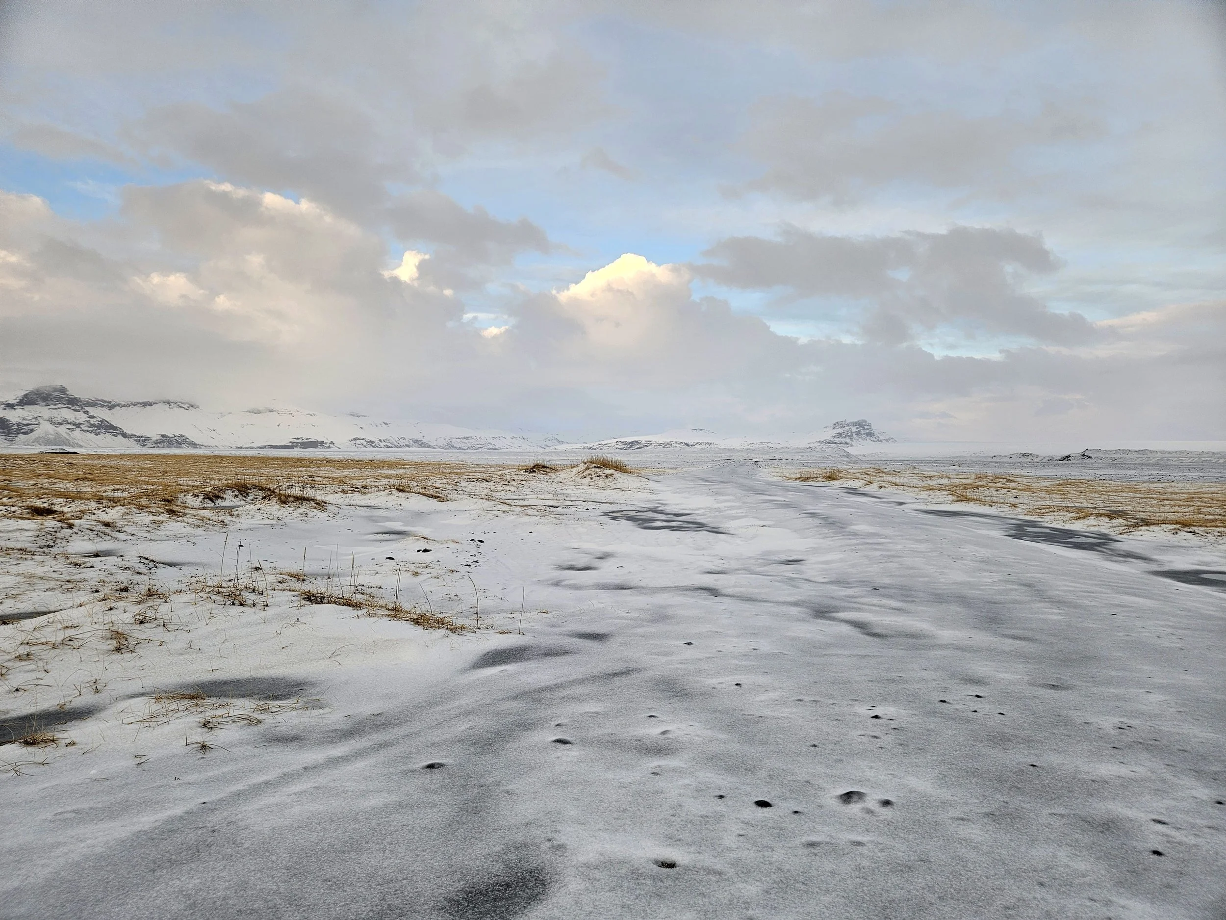 Snow-covered landscape with a cloudy sky, distant mountains, and footprints in the snow.
