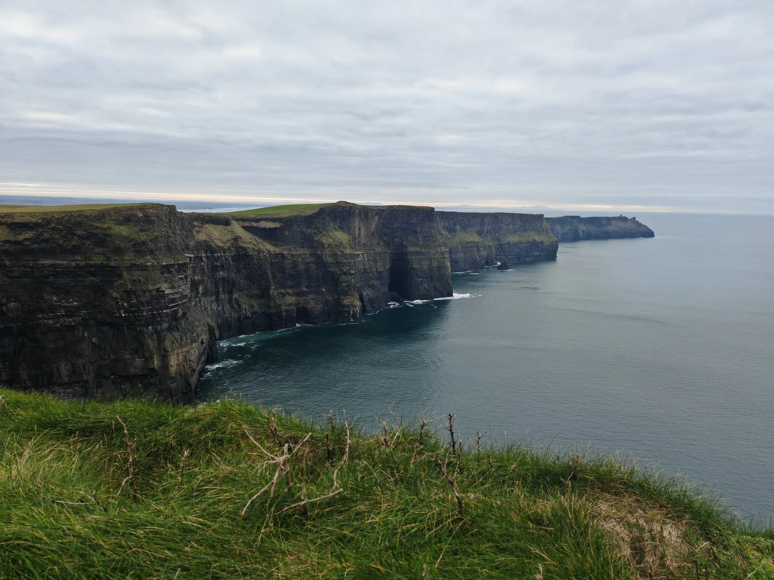 Cliffs along a coastline with grassy top and ocean below, under a cloudy sky.