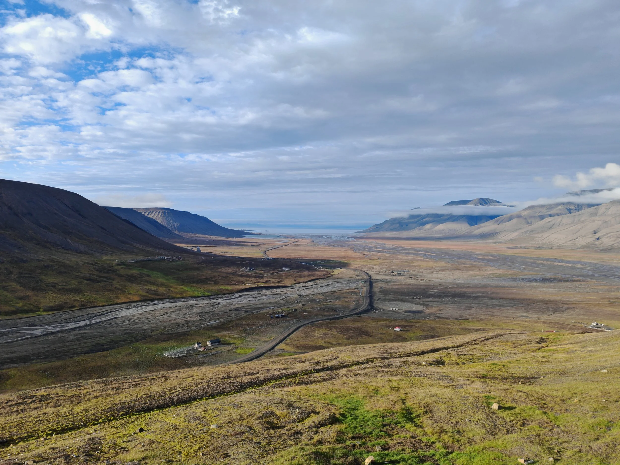 A vast landscape with rolling hills in shades of green and brown, a river valley in the distance, and clouds covering part of the sky over distant mountains.
