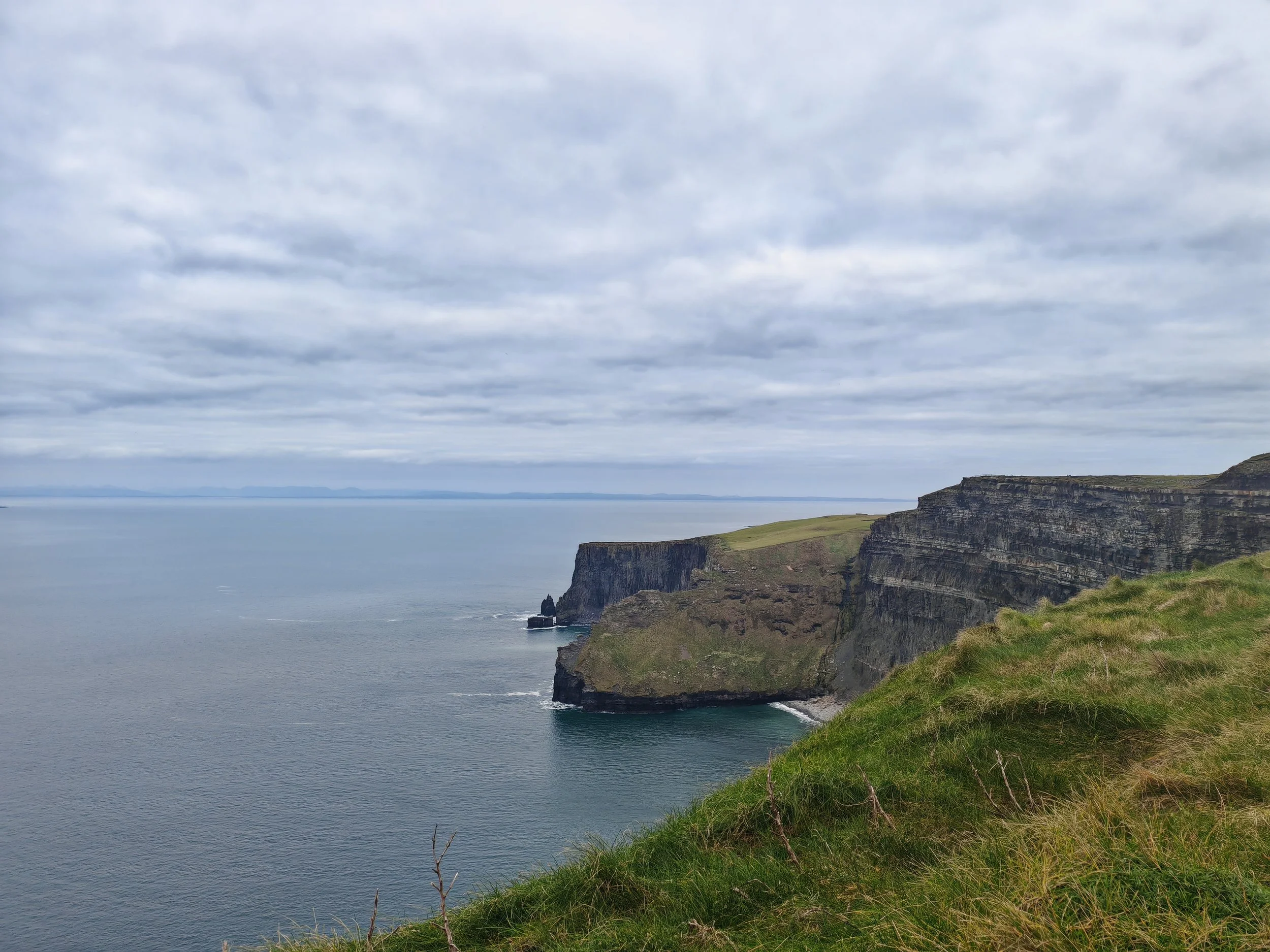 Cliffs over the ocean under a cloudy sky, with green grass at the edge of the cliff.