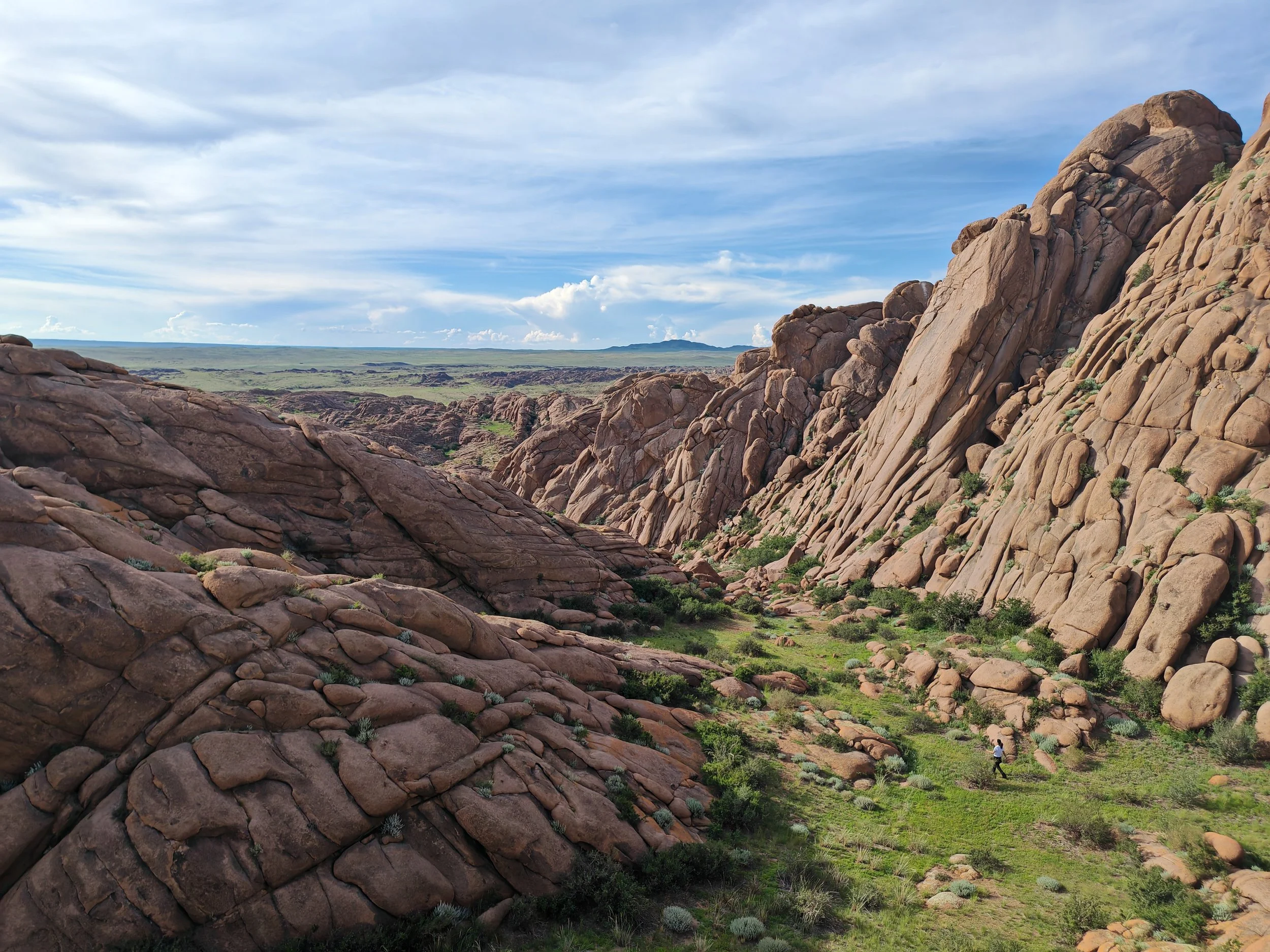 Scenic landscape of rocky terrain with large reddish-brown boulders and cliffs, green vegetation in the foreground, and an expansive sky with scattered clouds.