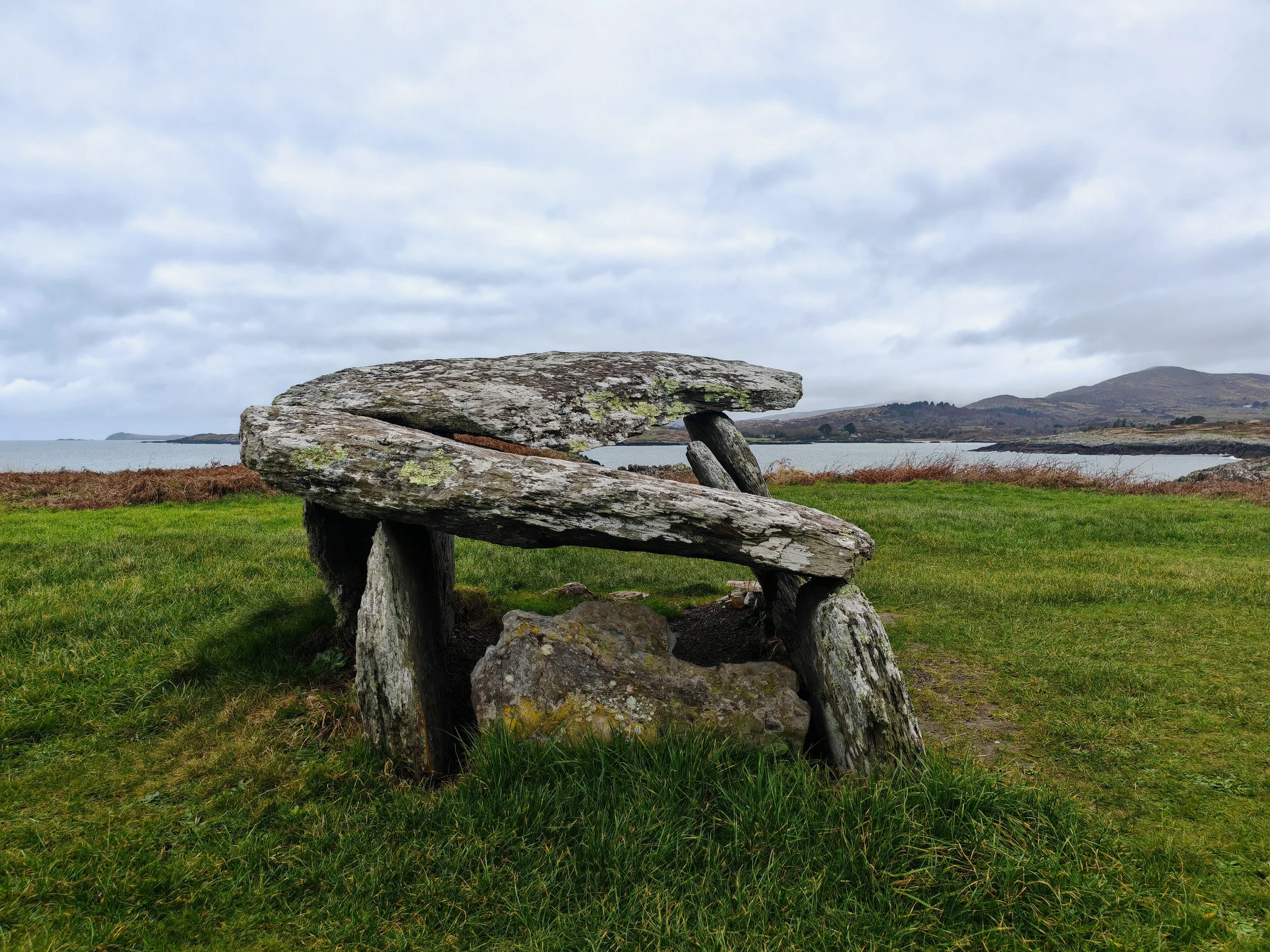 A large, flat rock balanced on smaller rocks in a grassy field near a body of water, with hills in the background and a cloudy sky overhead.