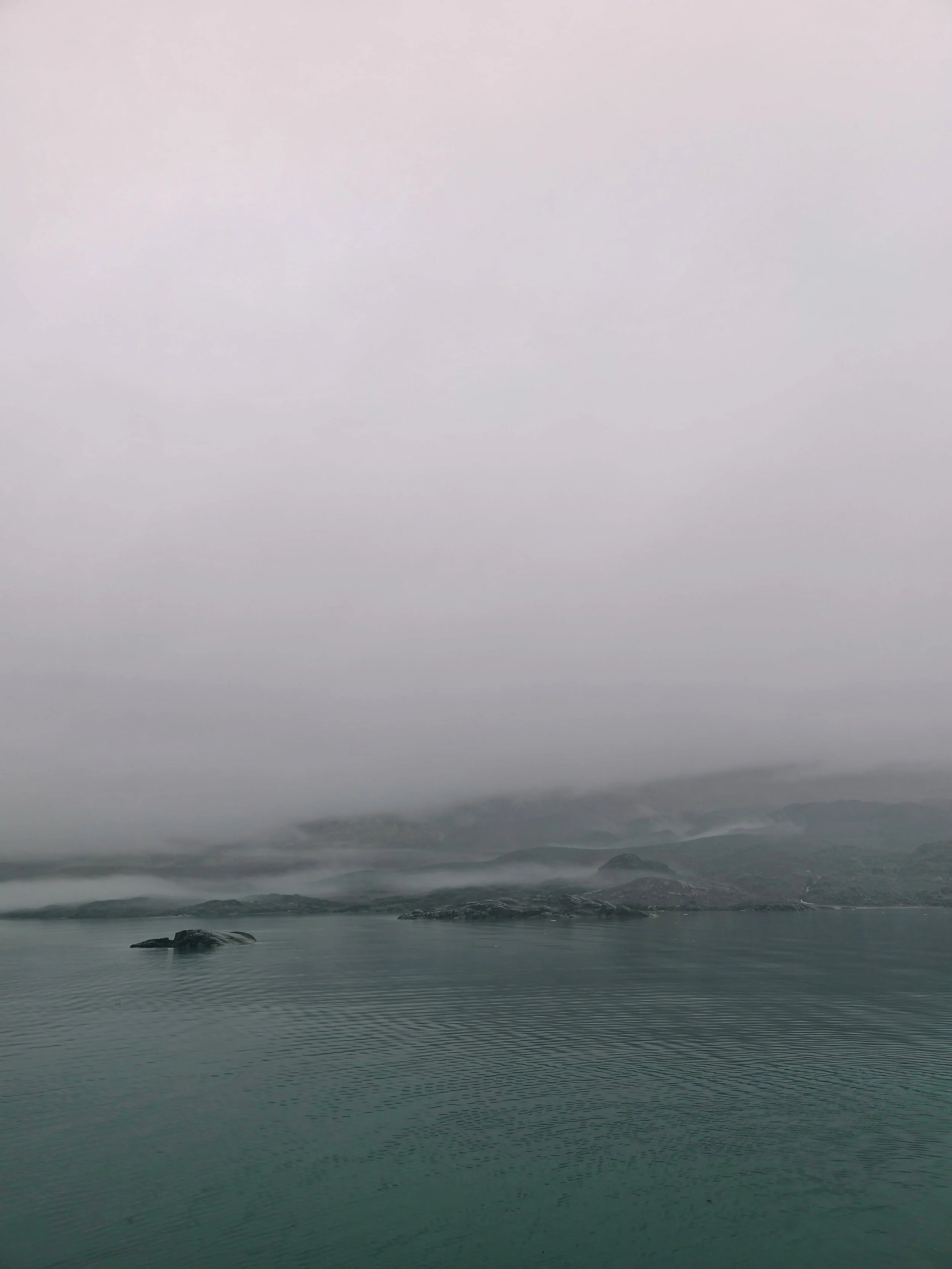 A foggy landscape of a body of water with some rocks and small islands, surrounded by misty hills or mountains in the background.