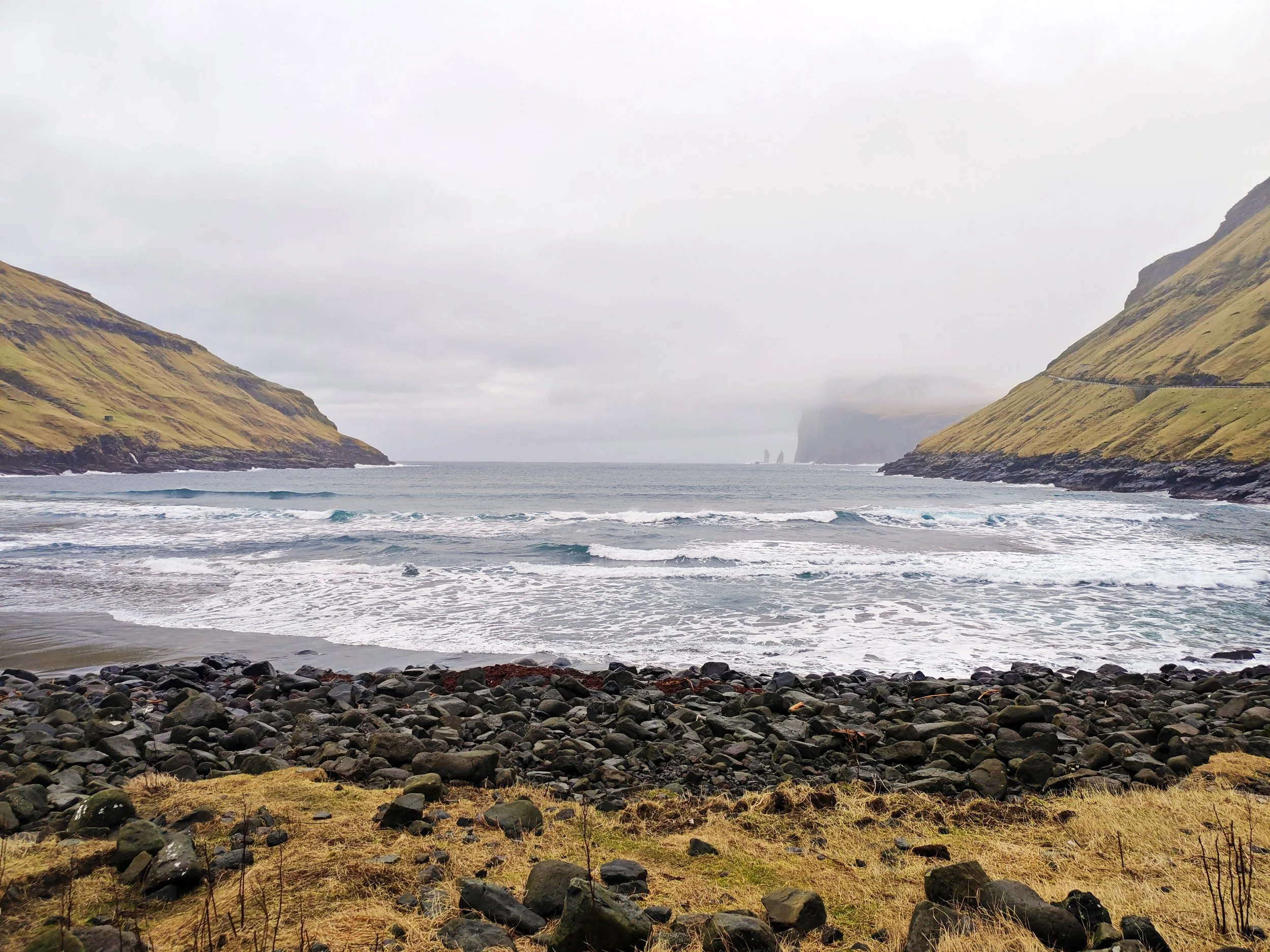 A rocky shoreline with dark stones in the foreground, green grassy hills on either side, and a body of water with small waves. Overcast sky with gray clouds.