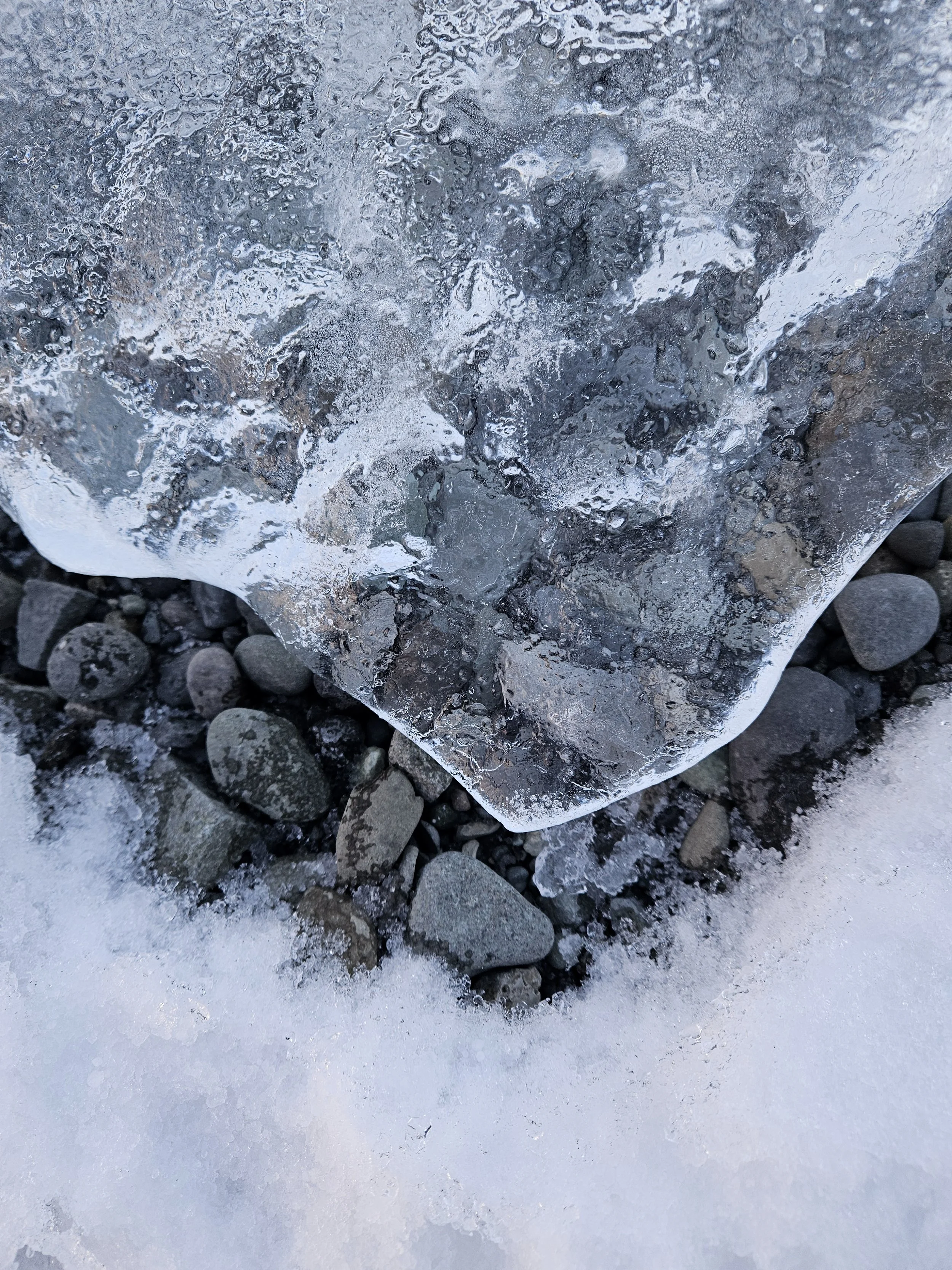 Close-up of a large, clear ice block on ice-covered ground surrounded by small rocks.