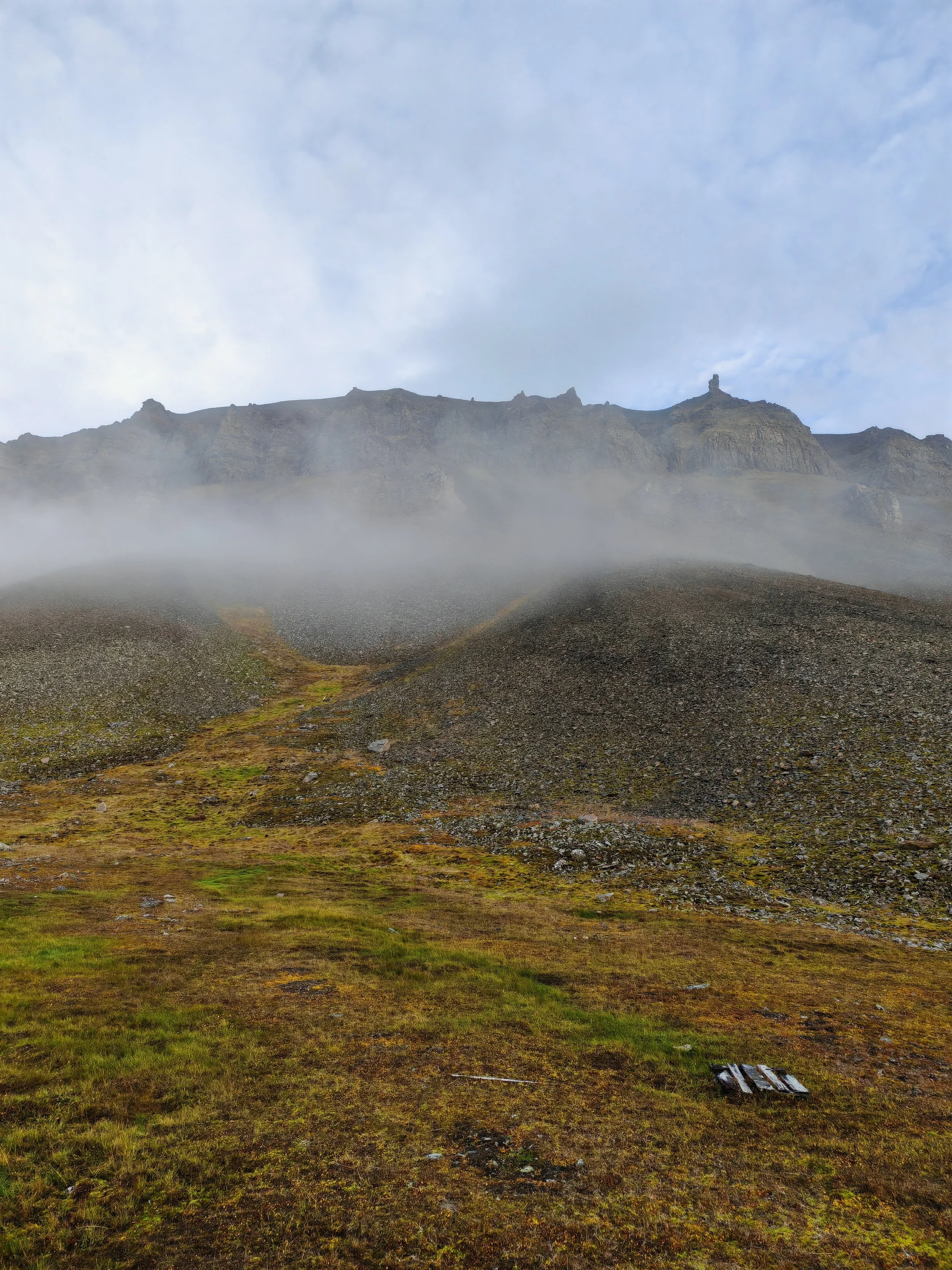 A landscape view of a mountain with rocky cliffs and some fog at the base, with patches of green and brown grass in the foreground, under a blue sky with scattered clouds.