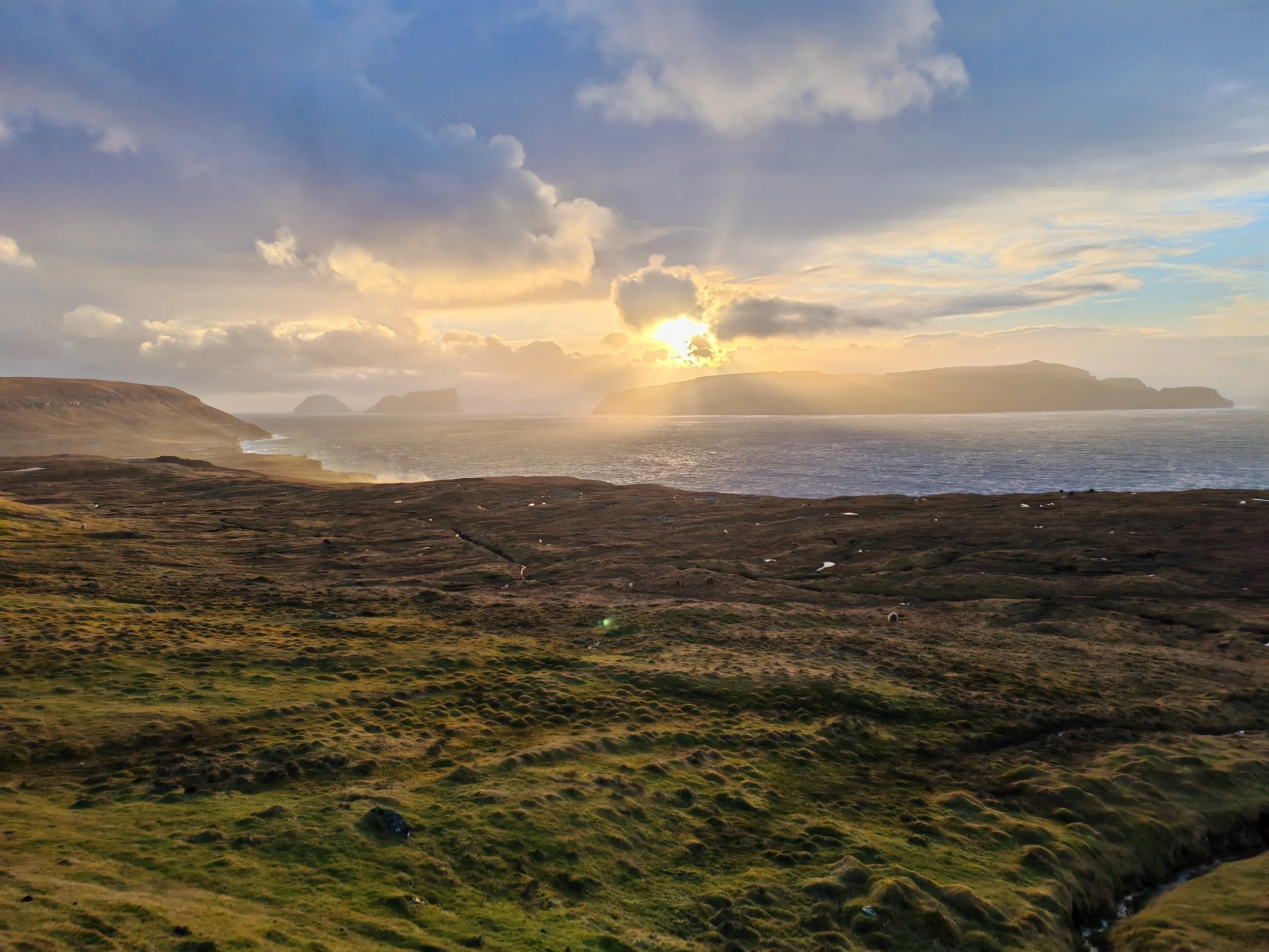 A scenic view of a coastline during sunset with cliffs, a body of water, cloudy sky, and sunlight breaking through clouds.