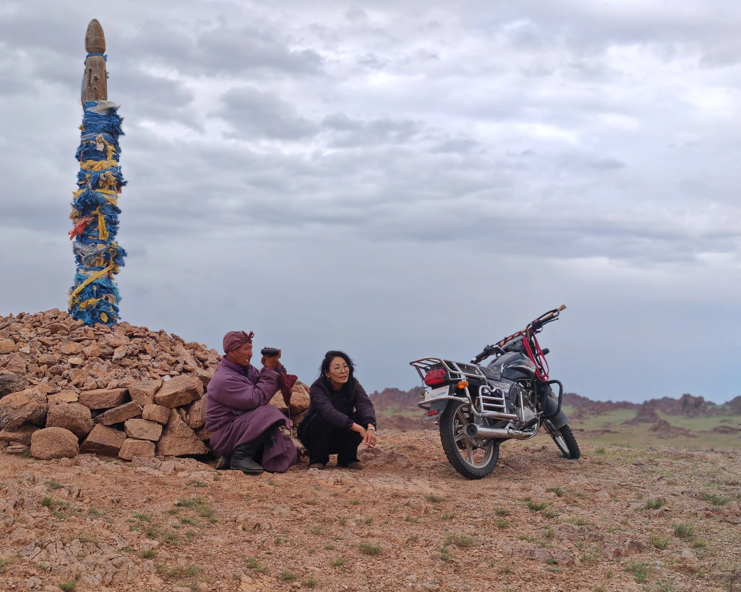 Two women sit on a rocky outdoor landscape, one woman takes a photo while the other looks into the distance, nearby a motorcycle is parked, and a colorful totem pole wrapped with blue, yellow, and white fabric stands in the background under a cloudy 