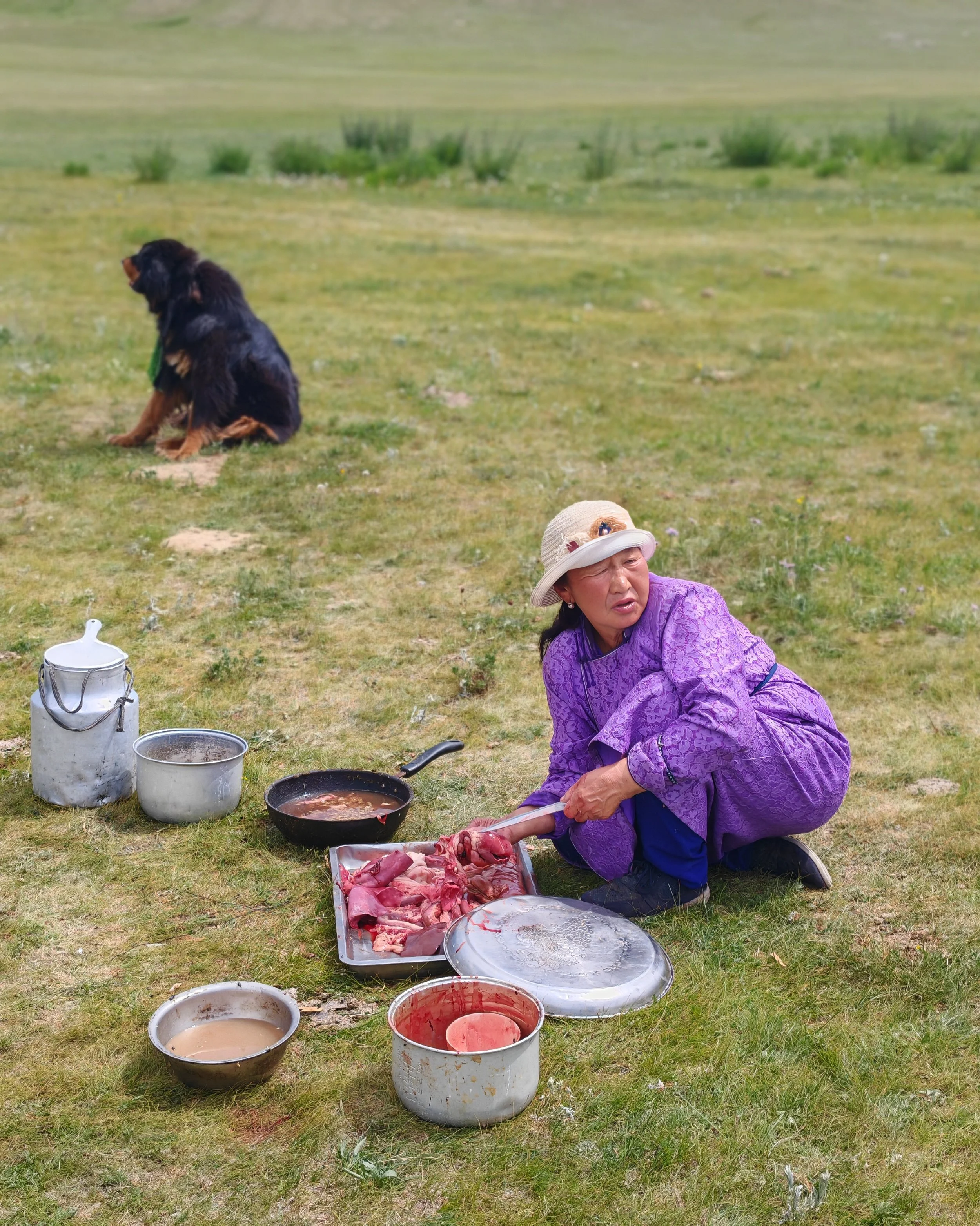 An elderly woman in a purple dress and sun hat kneels on grass, preparing raw meat with a knife on a tray, with cooking supplies including pots and buckets nearby, in an outdoor setting.