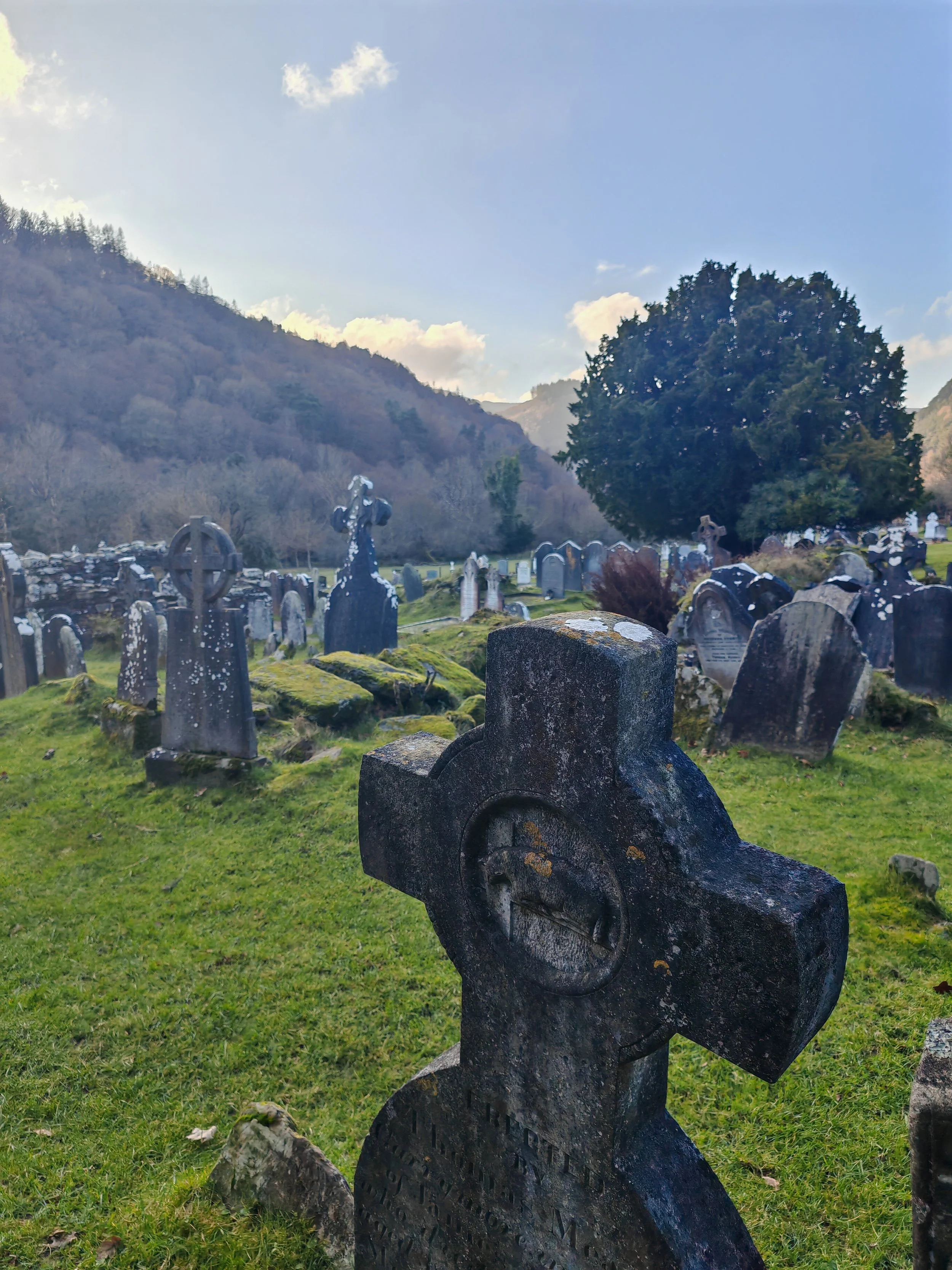 A graveyard with old tombstones and crosses, surrounded by a grassy area, with mountains and a blue sky with clouds in the background.