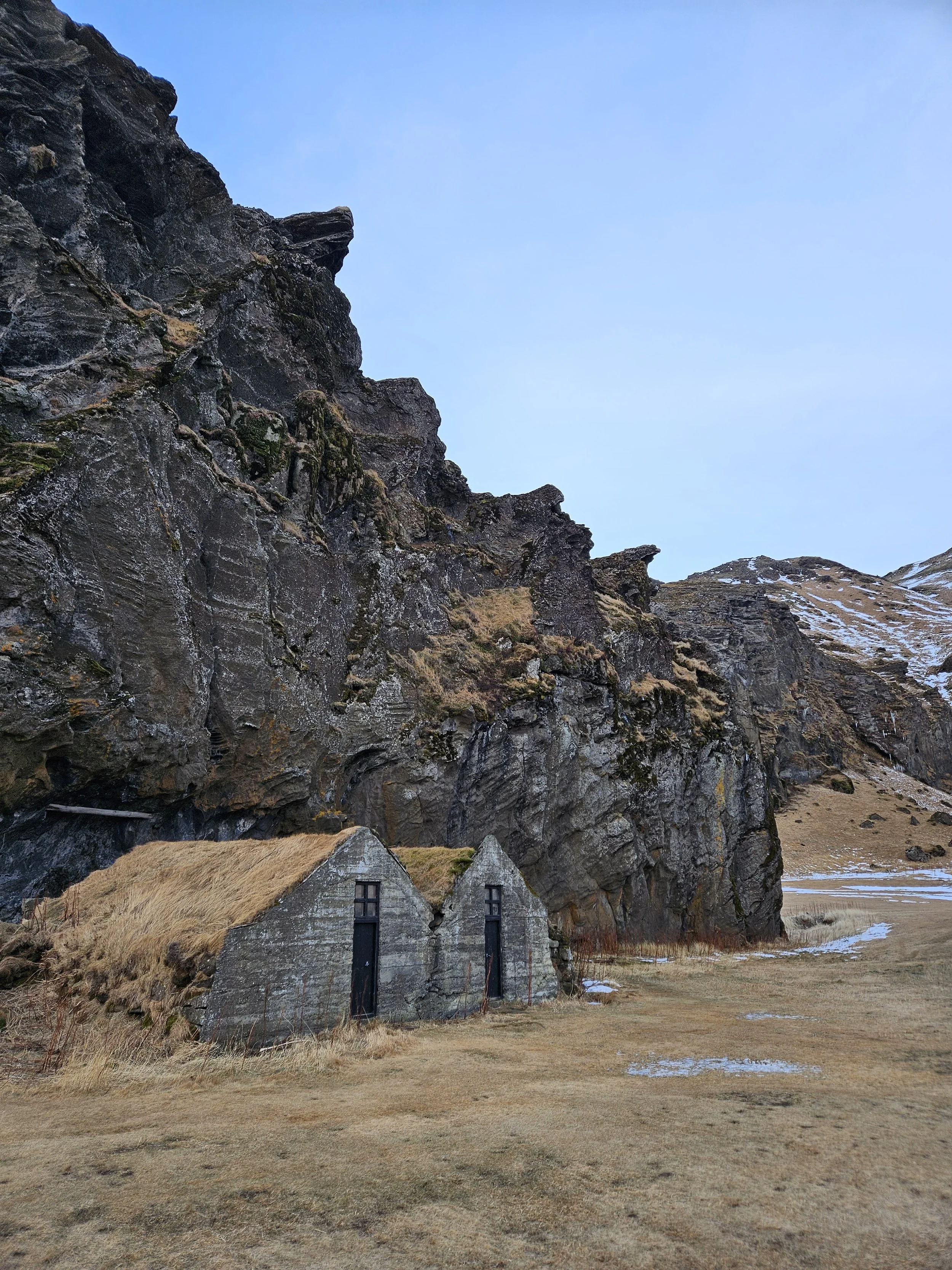 A rustic stone house with a thatched roof at the base of a rocky mountain with patches of snow, under a cloudy sky.