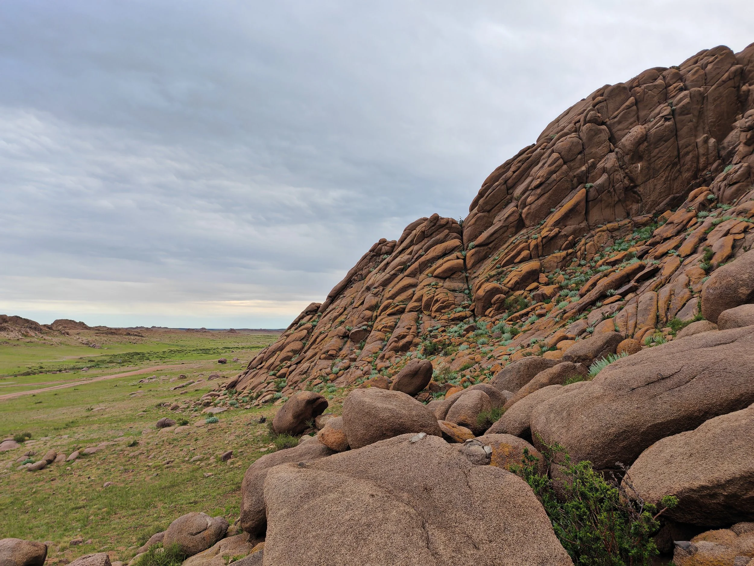 A rugged hillside with large boulder formations covered in sparse shrubbery, overlooking a flat grassy plain under a cloudy sky.