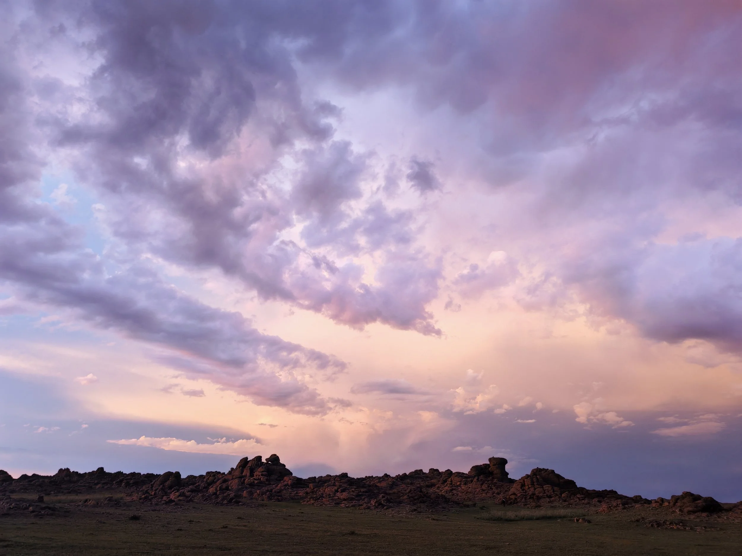 A rocky desert landscape with several large rocks and formations, under a colorful sky with purple, pink, and blue clouds at sunset.